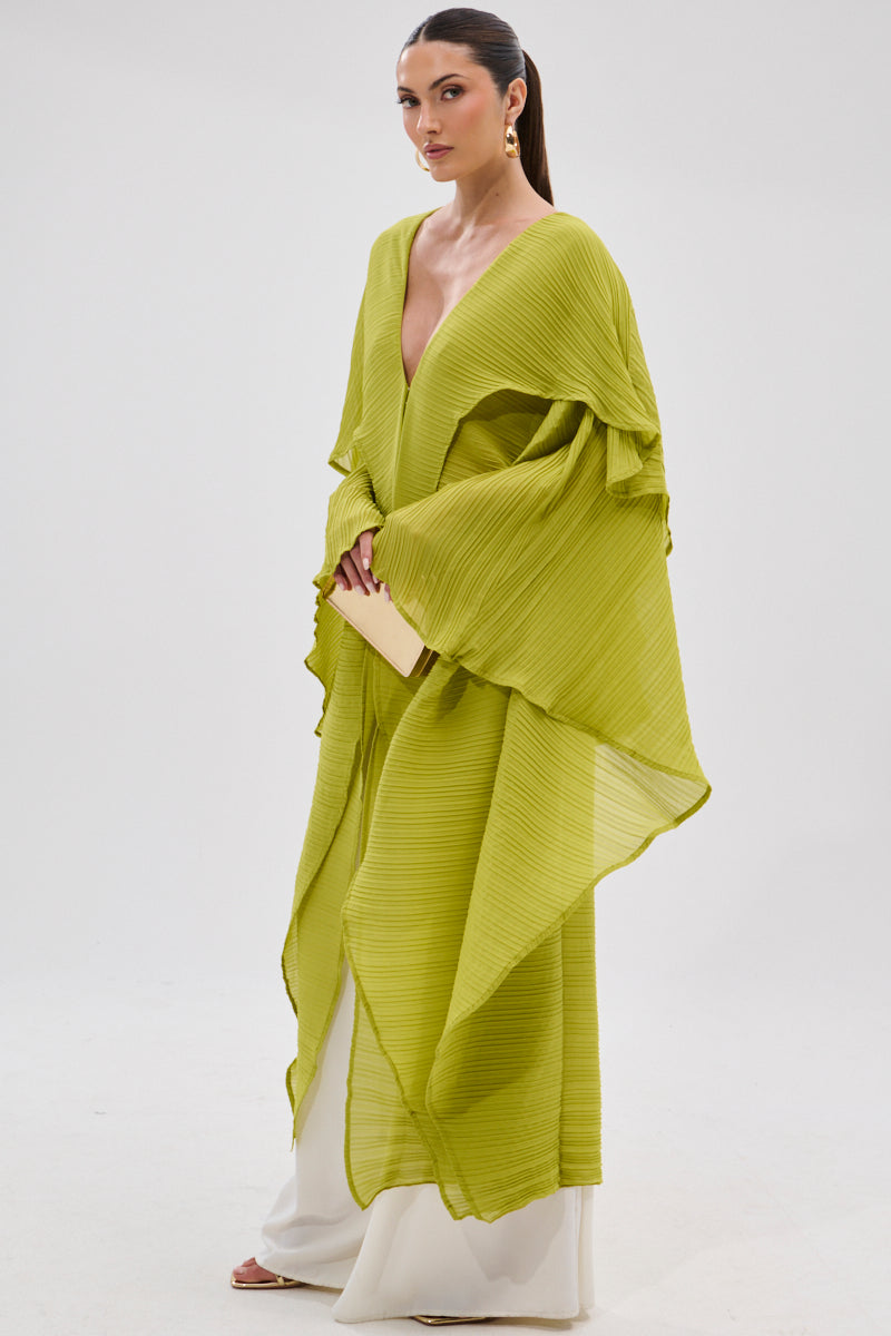 A woman stands in a studio wearing the FLY AWAY WITH ME BLOUSE IN CHARTREUSE with wide sleeves over white pants, styled with a sleek ponytail, gold earrings, and a beige clutch against a plain white background. 4