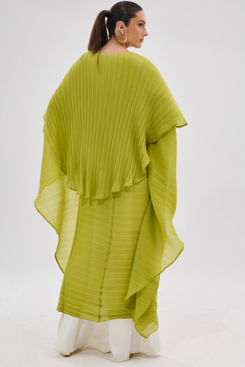 A woman with dark hair in a ponytail faces away, wearing the FLY AWAY WITH ME BLOUSE IN CHARTREUSE—a flowing, pleated longline poncho—over a white outfit. The background is plain and white. 6