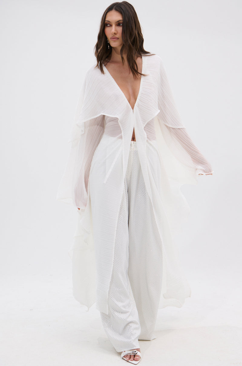 A woman in the FLY AWAY WITH ME BLOUSE IN WHITE—a sheer, flowing longline blouse with long sleeves—pairs it with wide-leg pants and white open-toe heels. She walks forward against a white background with loose wavy hair.