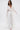 A woman in the FLY AWAY WITH ME BLOUSE IN WHITE—a sheer, flowing longline blouse with long sleeves—pairs it with wide-leg pants and white open-toe heels. She walks forward against a white background with loose wavy hair.