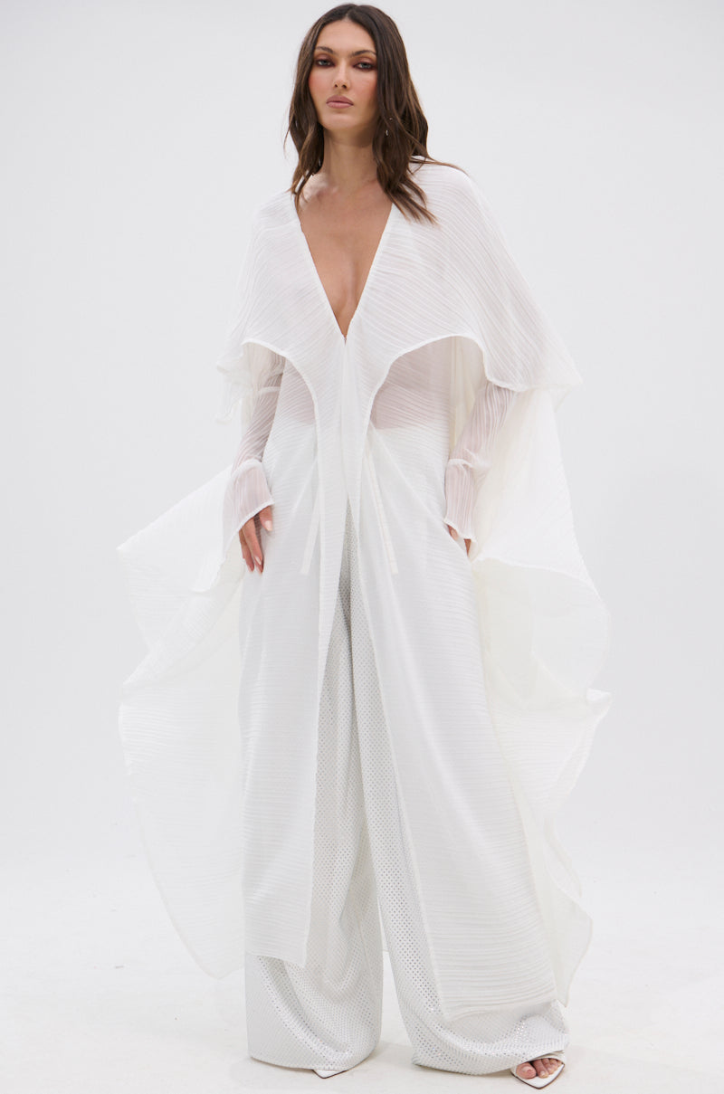 A woman wears the FLY AWAY WITH ME BLOUSE IN WHITE, a flowing, sheer top with wide pleated sleeves and a deep V-neck, paired with loose textured pants for an elegant, ethereal look against a simple background.