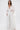 A woman wears the FLY AWAY WITH ME BLOUSE IN WHITE, a flowing, sheer top with wide pleated sleeves and a deep V-neck, paired with loose textured pants for an elegant, ethereal look against a simple background.