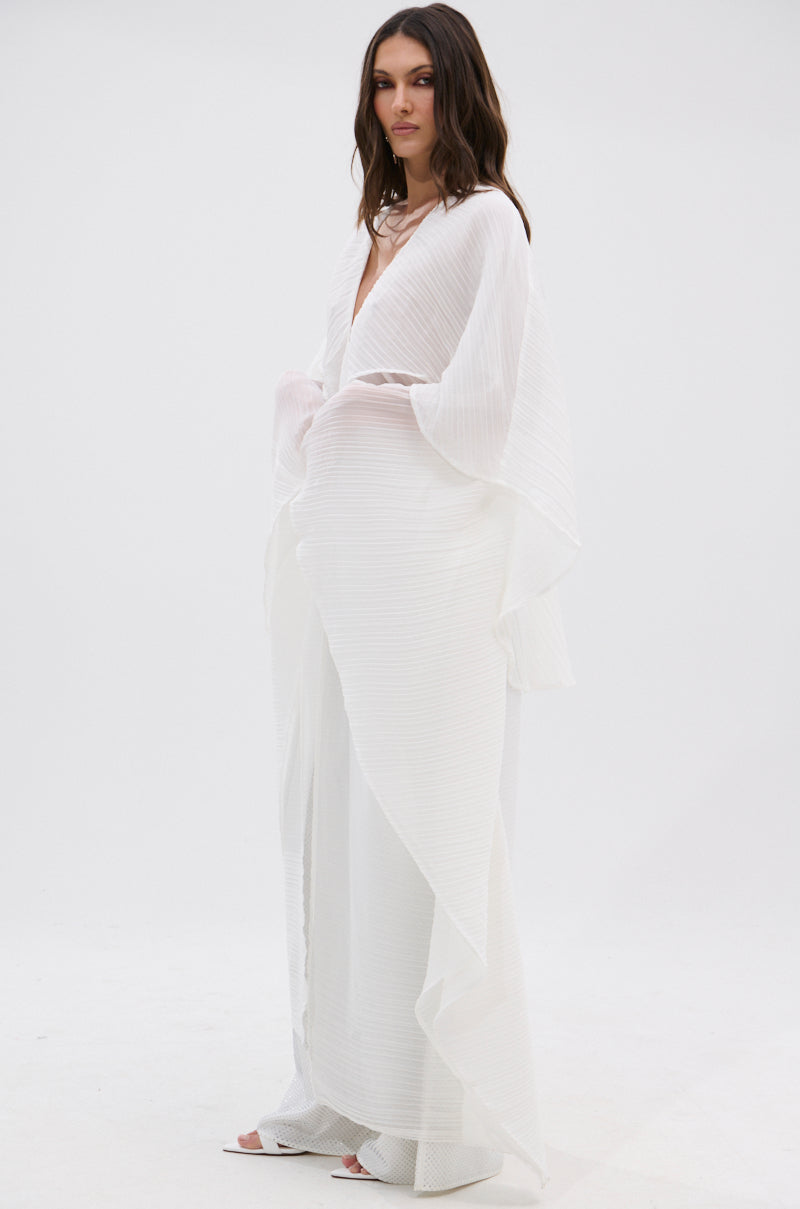 A woman with long, wavy brown hair and a calm expression wears the FLY AWAY WITH ME BLOUSE IN WHITE—a sheer, flowing top with wide draped sleeves and subtle vertical stripes—standing against a plain white background.