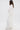 A woman with long, wavy brown hair stands sideways against a plain white background wearing the FLY AWAY WITH ME BLOUSE IN WHITE—featuring layered, draped fabric and a flowing, textured design.