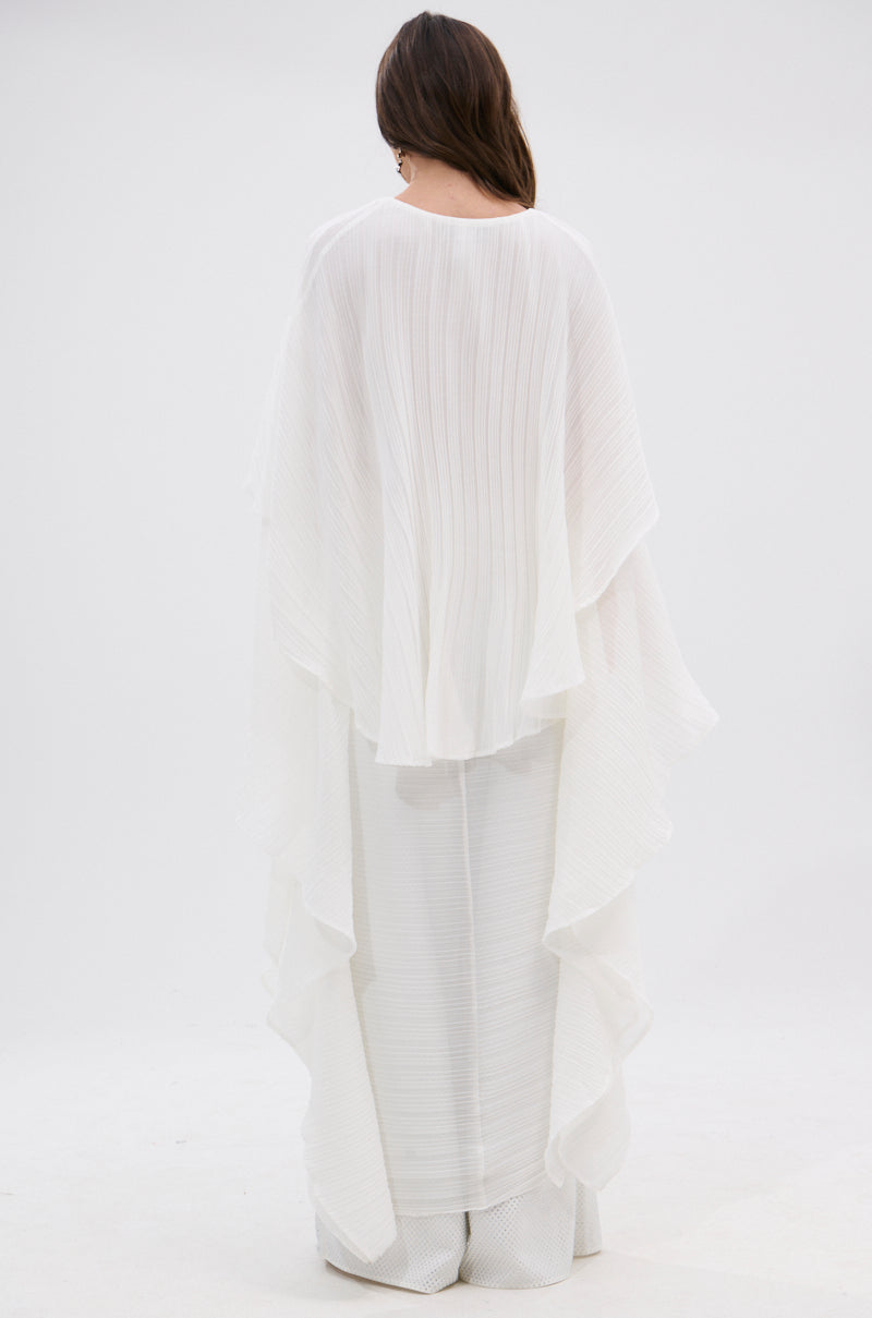 A woman with long brown hair, seen from behind, wears the FLY AWAY WITH ME BLOUSE IN WHITE—a longline blouse with pleated, cape-like details—paired with wide-leg pants against a plain white background.