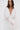 A woman with long brown hair wears the FLY AWAY WITH ME BLOUSE IN WHITE—a sheer, flowing pleated longline blouse with wide sleeves and a deep V-neck—standing against a plain light background.