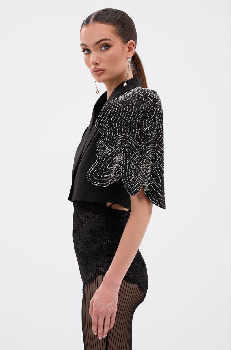 A woman stands in profile wearing the JUST WATCH EMBELLISHED CAPE, a black cropped blazer with rhinestone details on one shoulder, styled with lace high-waisted bottoms, striped sheer tights, slicked-back hair, and dangling earrings. 2
