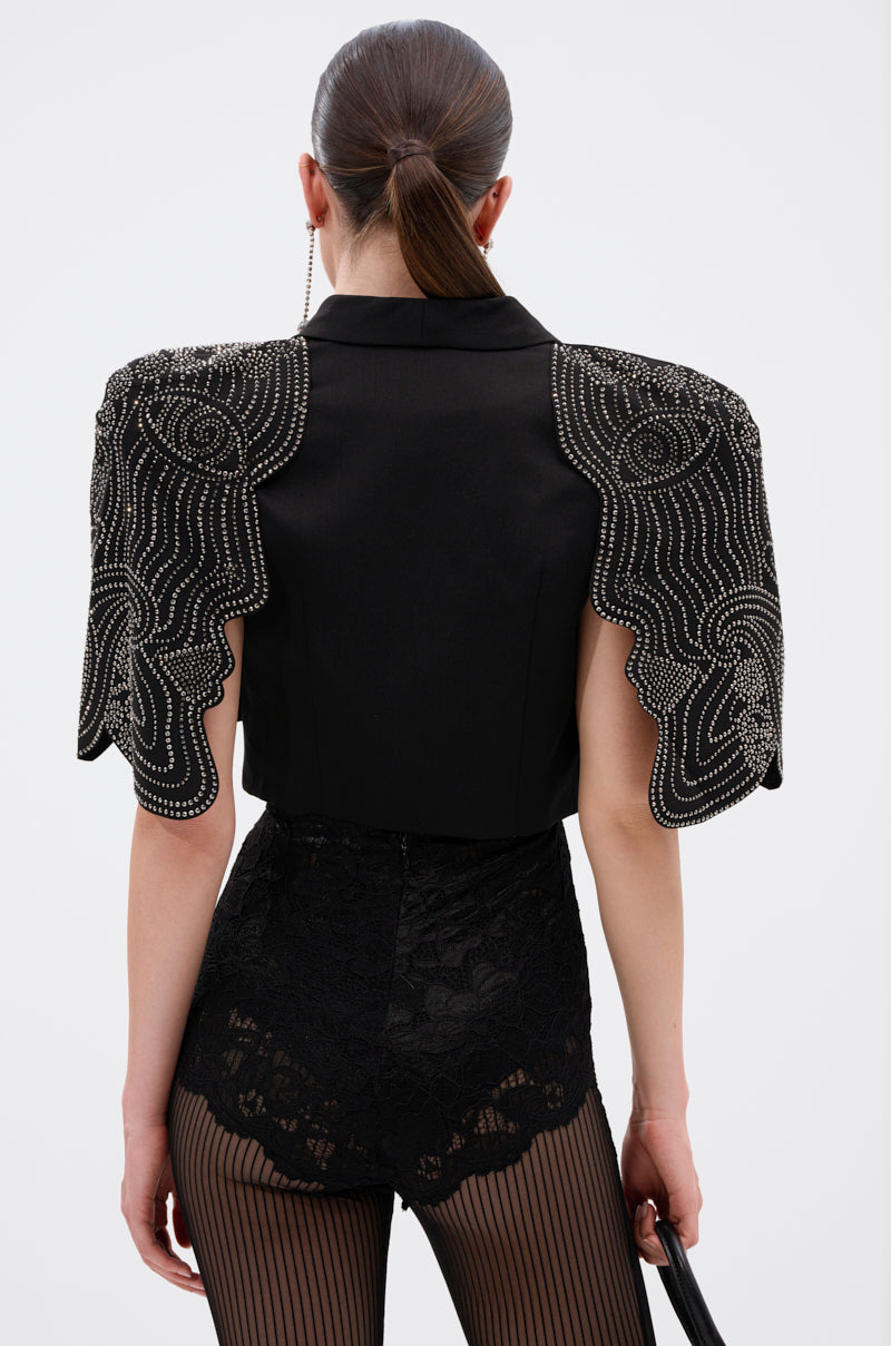 A woman with straight, dark hair pulled back is photographed from the back wearing the JUST WATCH EMBELLISHED CAPE, featuring rhinestone wing-like shoulders, paired with black lace shorts over sheer, vertically striped tights. 3