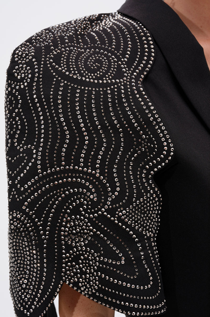 A close-up of the JUST WATCH EMBELLISHED CAPE, a black cropped blazer with dramatic shoulders, adorned with intricate silver studs in swirling geometric patterns. 6