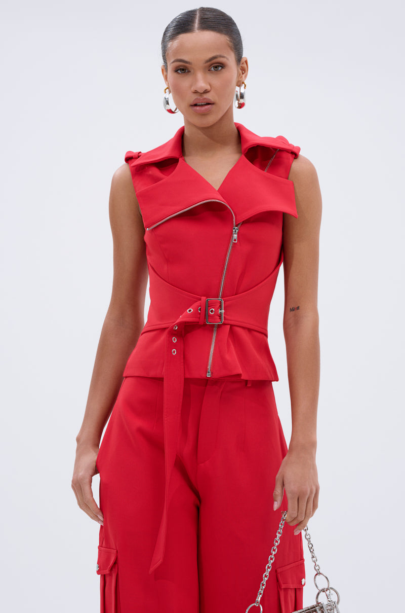 A woman wears the LAURA MOTO VEST, a bold red AKIRA Label sleeveless vest with a wide collar, front zipper, and belted waist, paired with matching high-waisted red pants. She holds a silver chain handbag and has her hair tied back. 0
