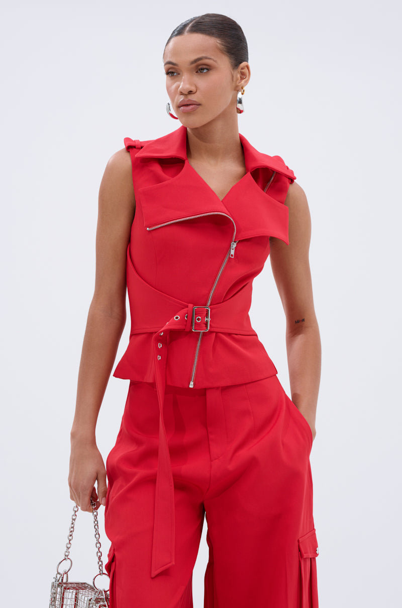 Wearing the LAURA MOTO VEST in red with an asymmetrical zipper, a woman pairs it with matching wide-leg pants and holds a small bag. Her hair is pulled back as she poses against a plain background, looking to the side. 2