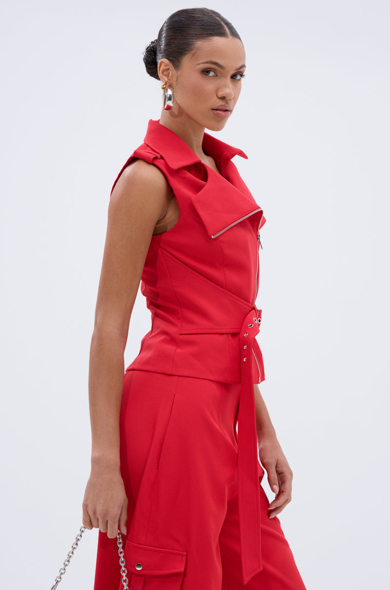A woman with her hair in a bun wears a sleeveless, belted red jumpsuit styled with the LAURA MOTO VEST—a tailored, wide-collared piece with zipper detail—holding a chain strap accessory and posing against a plain light background. 3