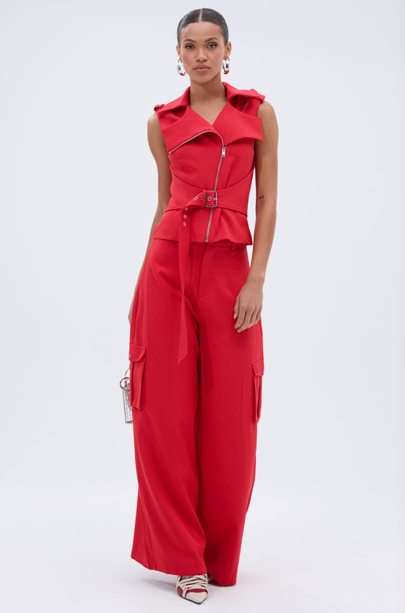 A woman wears the LAURA MOTO VEST, a bold red sleeveless piece with an asymmetrical zipper and belted waist, styled with wide-leg cargo pants, strappy heels, and hoop earrings against a white background. 6