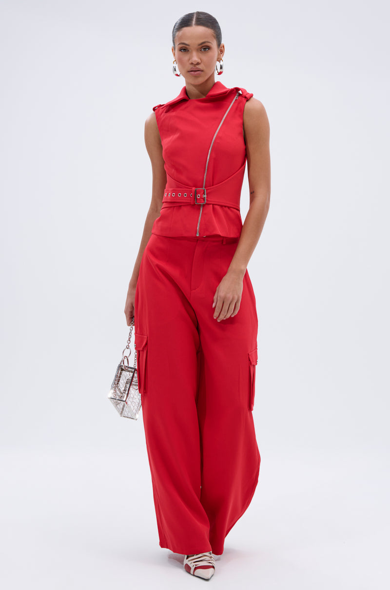 A woman models the LAURA MOTO VEST, a bright red sleeveless design with an asymmetrical zipper, paired with matching wide-leg pants. She carries a small handbag, wears white strappy heels, and accessorizes with large hoop earrings. 7