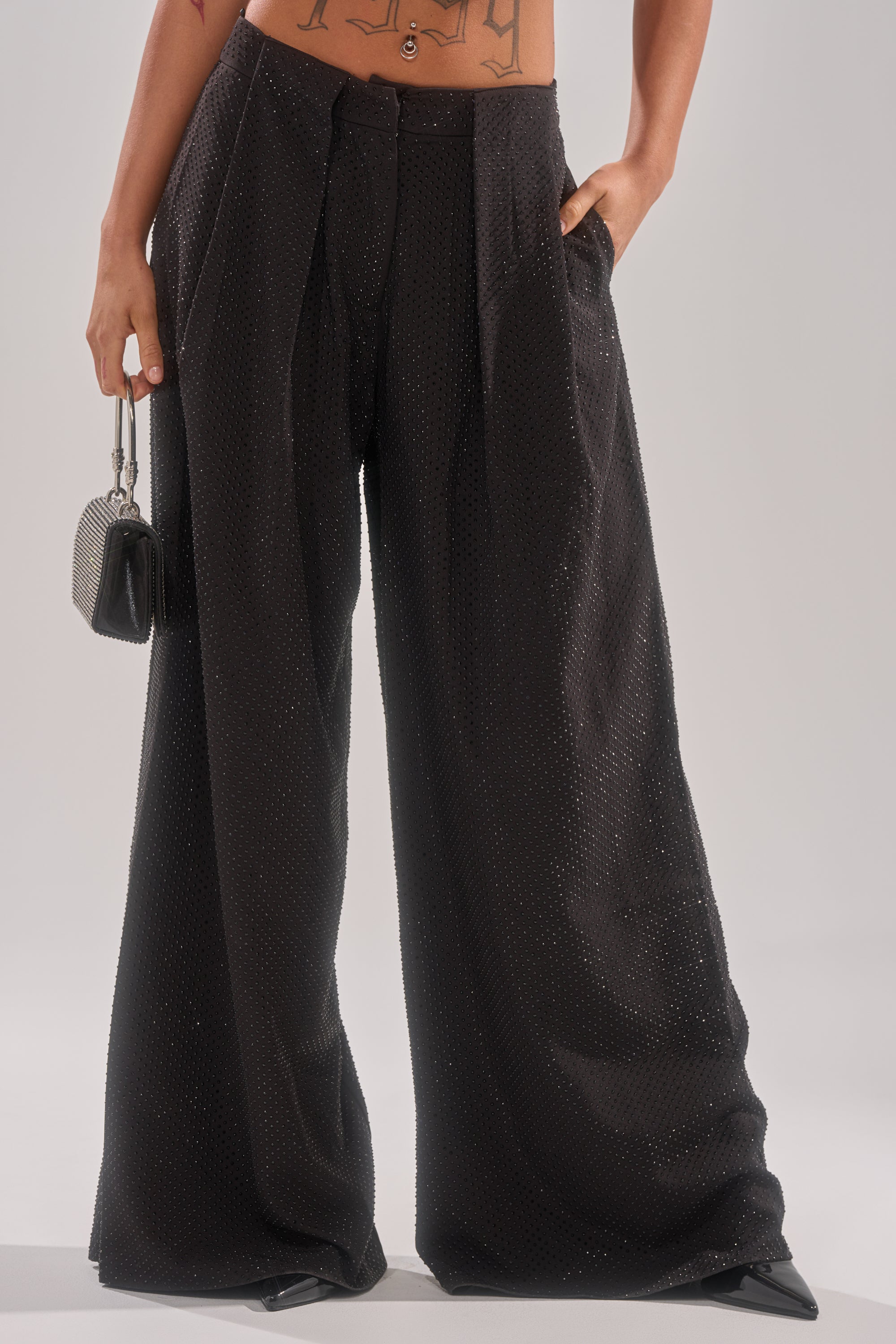 Wearing high-waisted SPARKLE QUEEN TROUSER with rhinestone details, a cropped top exposing midriff tattoos, paired with pointed black shoes and a small silver purse against a plain background. 2