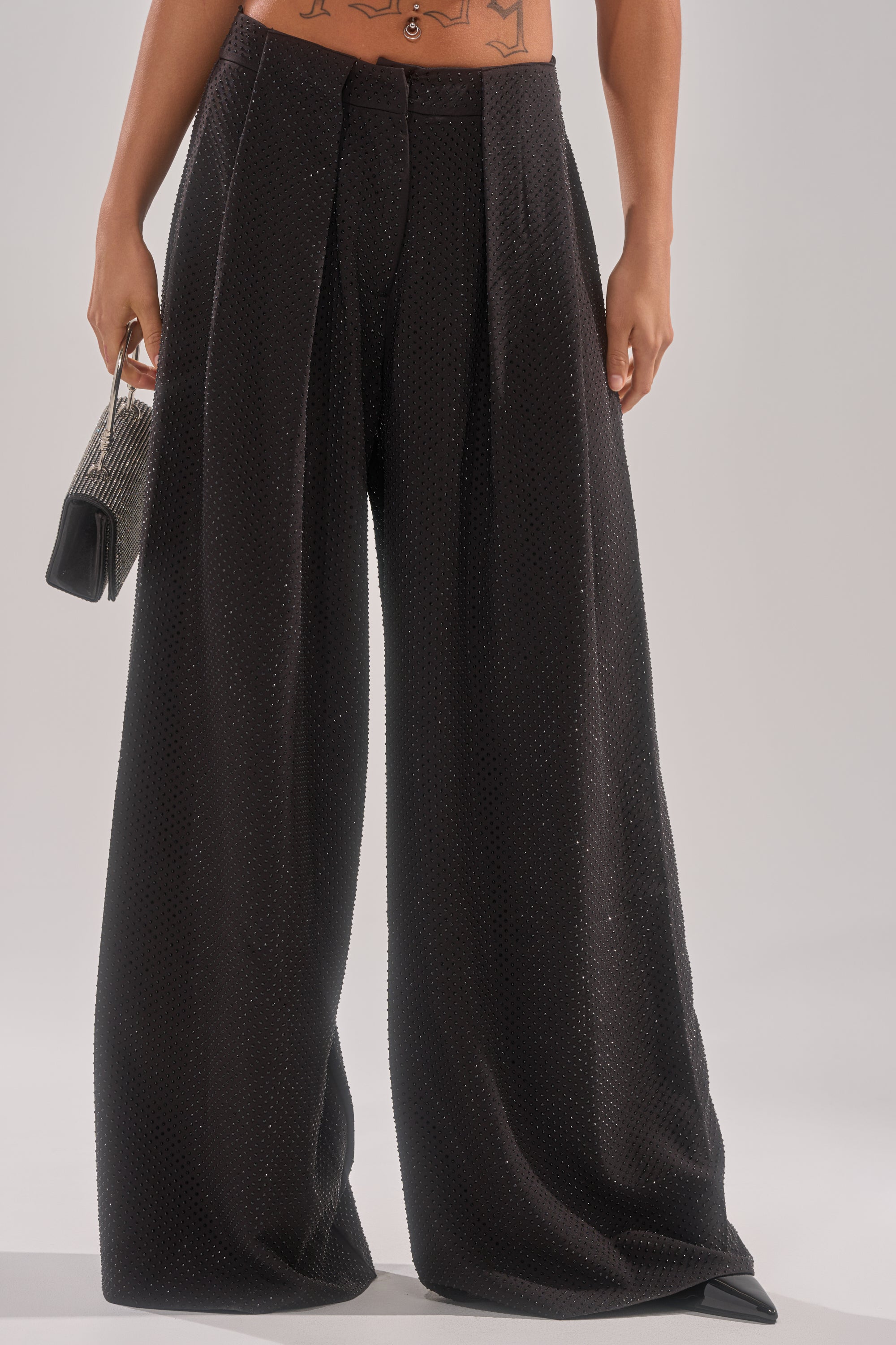 Wearing the SPARKLE QUEEN TROUSER—high-waisted, wide-leg trousers with subtle texture—paired with a small metallic handbag. The cropped image shows part of the midriff and a tattoo. 0