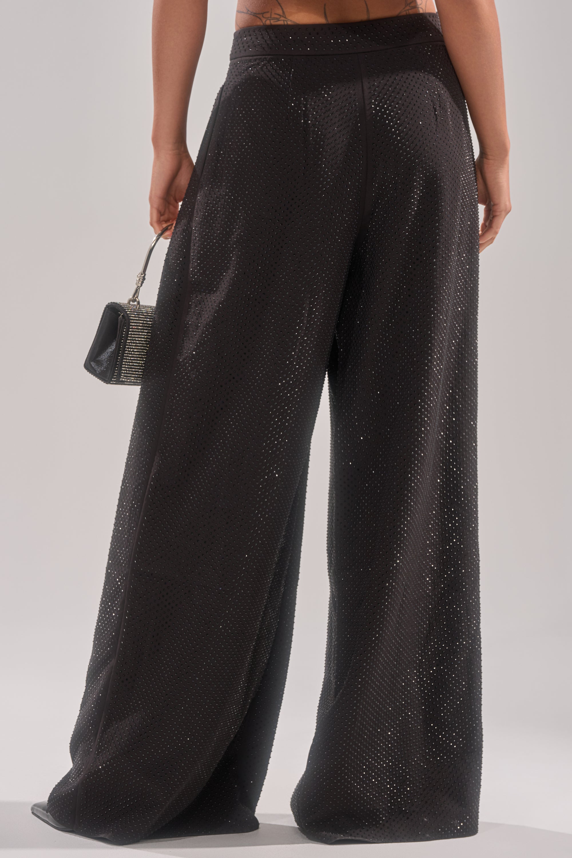 A person stands facing away in SPARKLE QUEEN TROUSER, wide-leg black sparkle pants, holding a small textured black handbag against a plain light background. 4