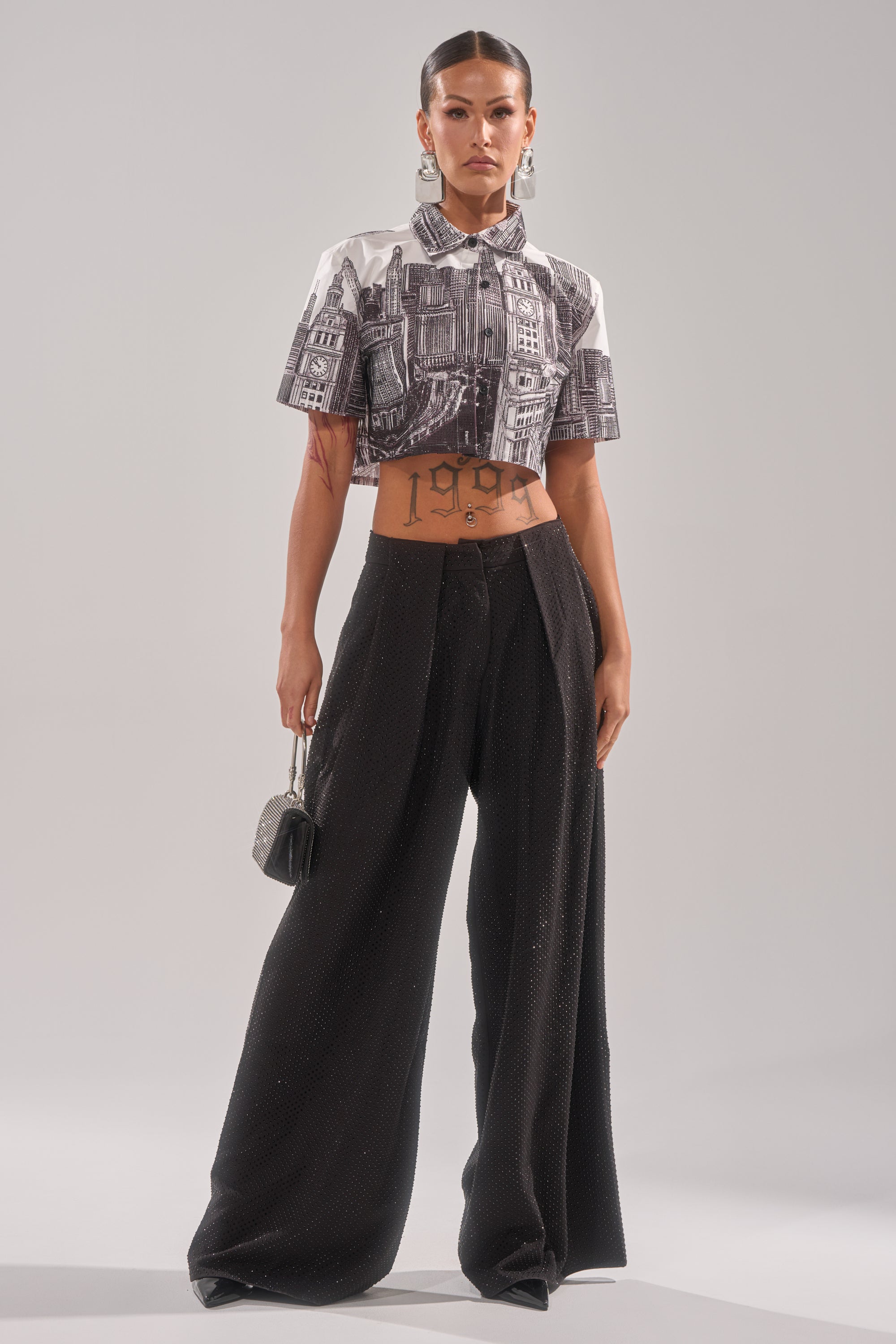 Wearing the SPARKLE QUEEN TROUSER, a person stands confidently in wide-leg black pants and a cropped cityscape-print shirt, accessorized with statement earrings, a small handbag, and sleek hair. 5