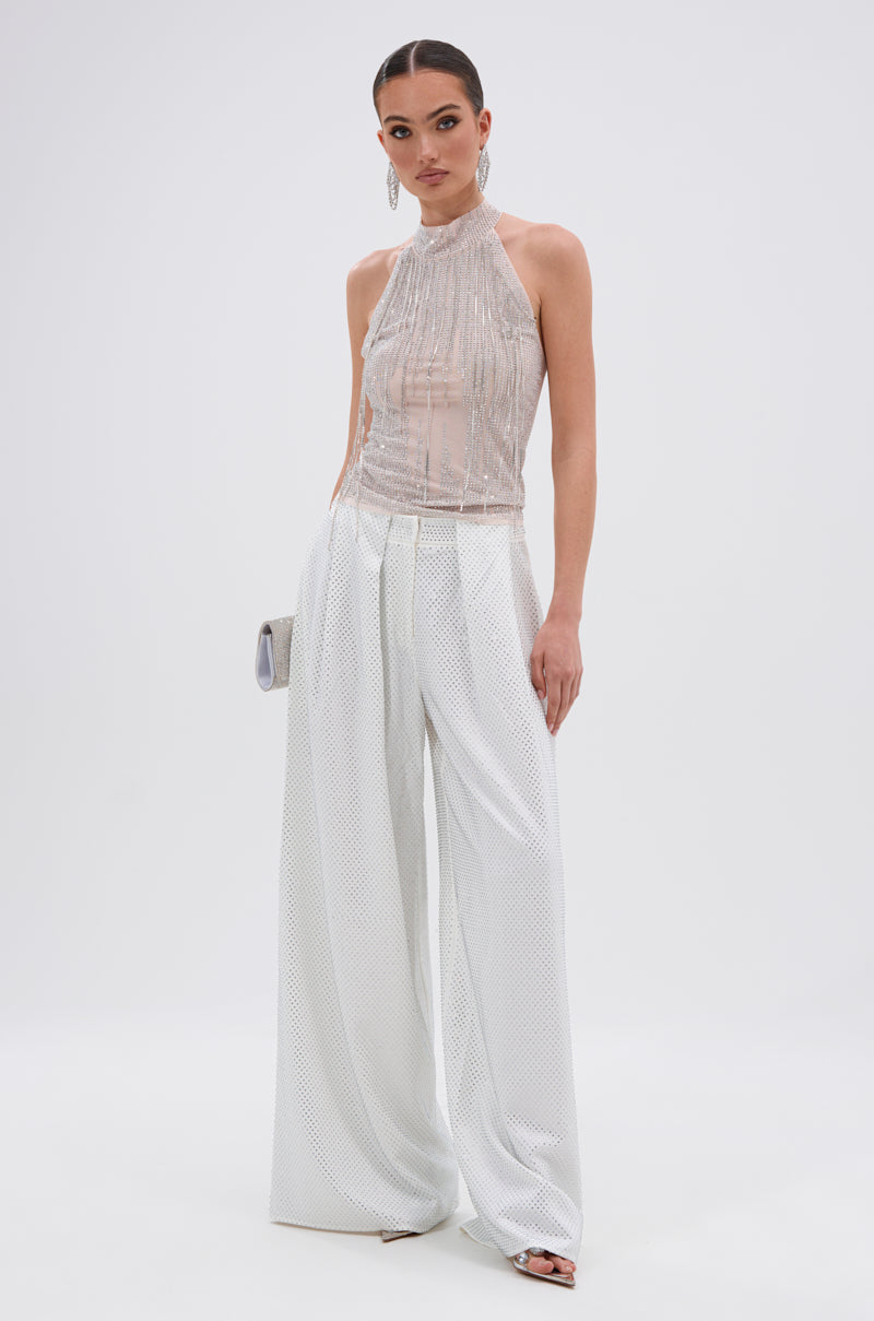 A woman with slicked-back hair stands against a white background, wearing a sleeveless sheer top and the SPARKLE QUEEN TROUSER with a wide-leg silhouette. She accessorizes with open-toe heels and a small silver clutch.