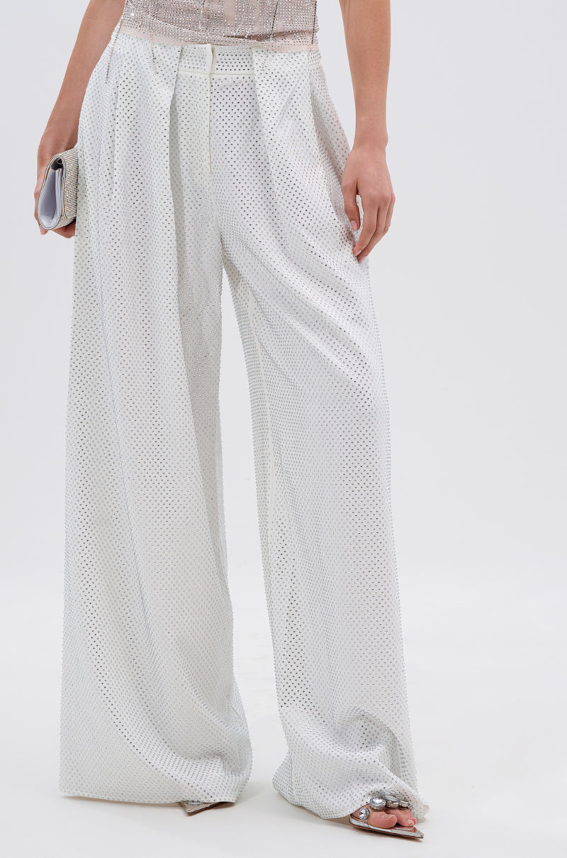 A woman wears the SPARKLE QUEEN TROUSER, featuring a loose white wide-leg silhouette with subtle mesh texture, paired with a silver clutch and rhinestone-embellished open-toe sandals.