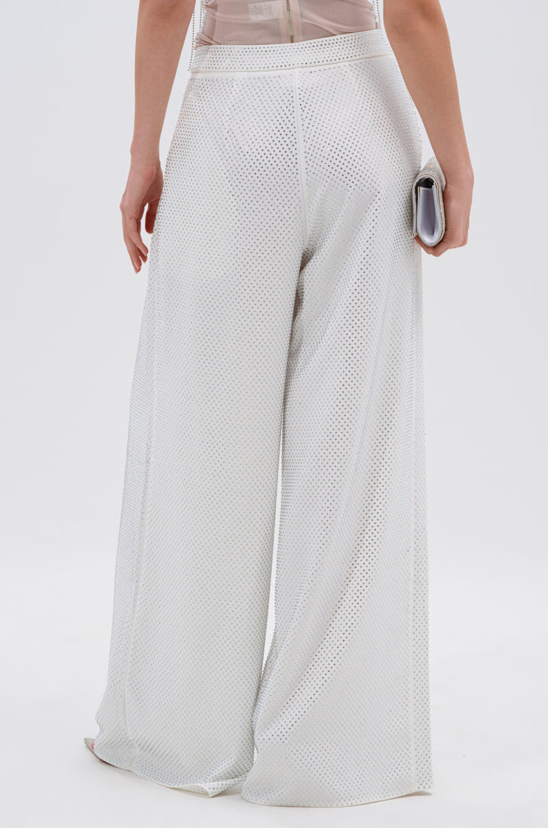 A person faces away, wearing the SPARKLE QUEEN TROUSER—white, perforated, and wide-leg—while holding a small clutch against a plain white background.