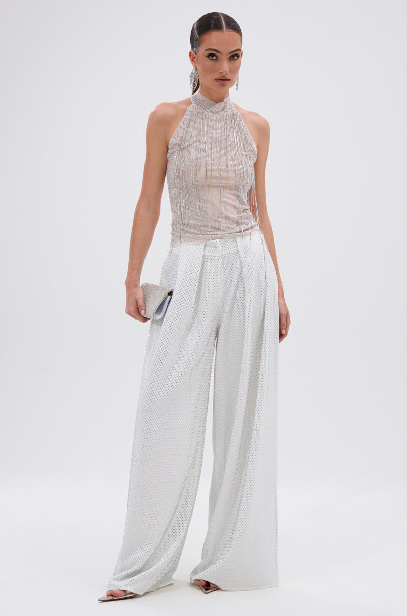 A woman poses against a white background in a sleeveless, high-neck shimmery top and SPARKLE QUEEN TROUSER with wide-leg silhouette, holding a small clutch and wearing her hair slicked back.