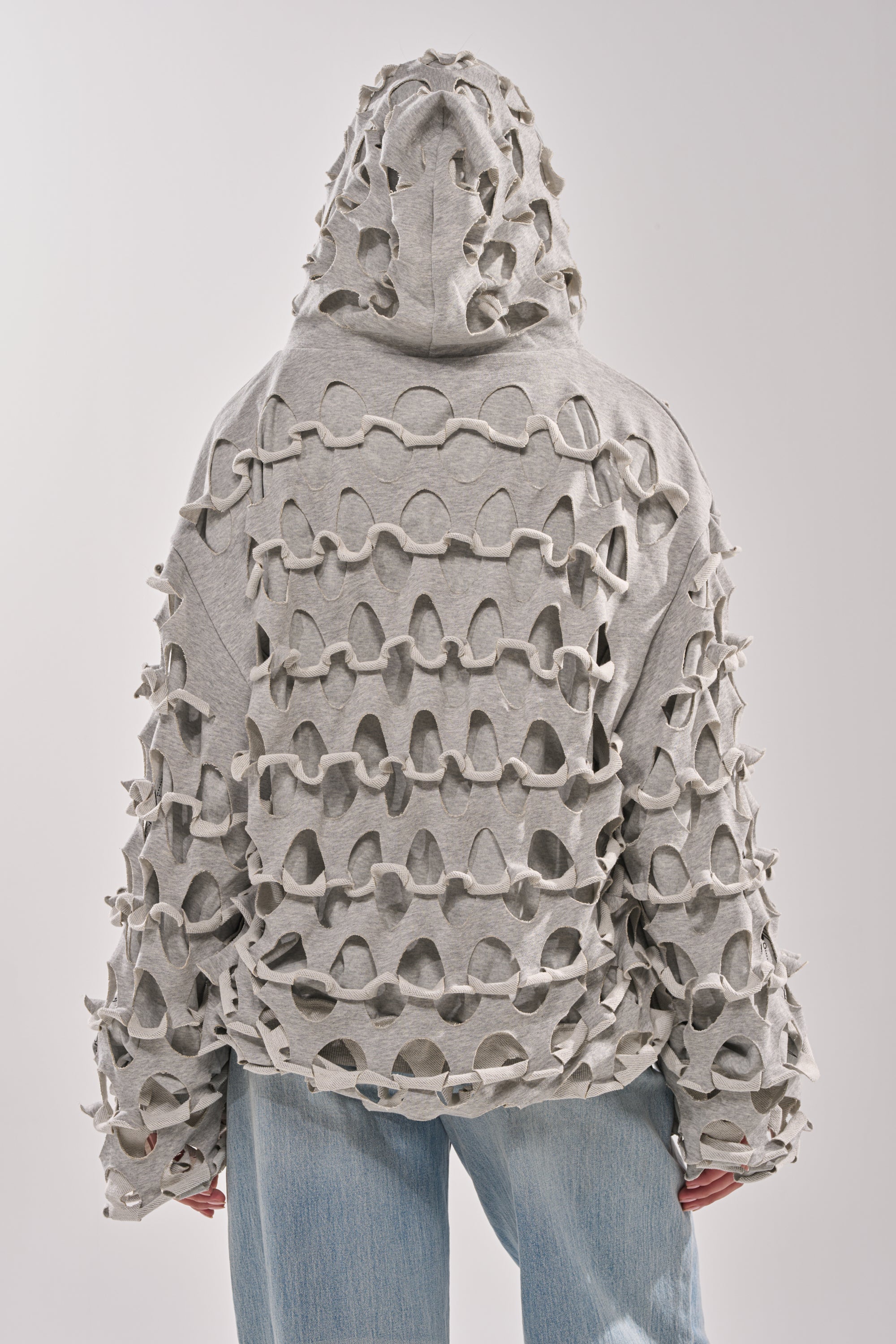 A person is shown from the back against a plain light background, wearing the EPIC REVERSIBLE ZIP UP SWEATSHIRT in light gray with an oversized hood, intricate cutout pattern, and raised loops, paired with light blue jeans. 11