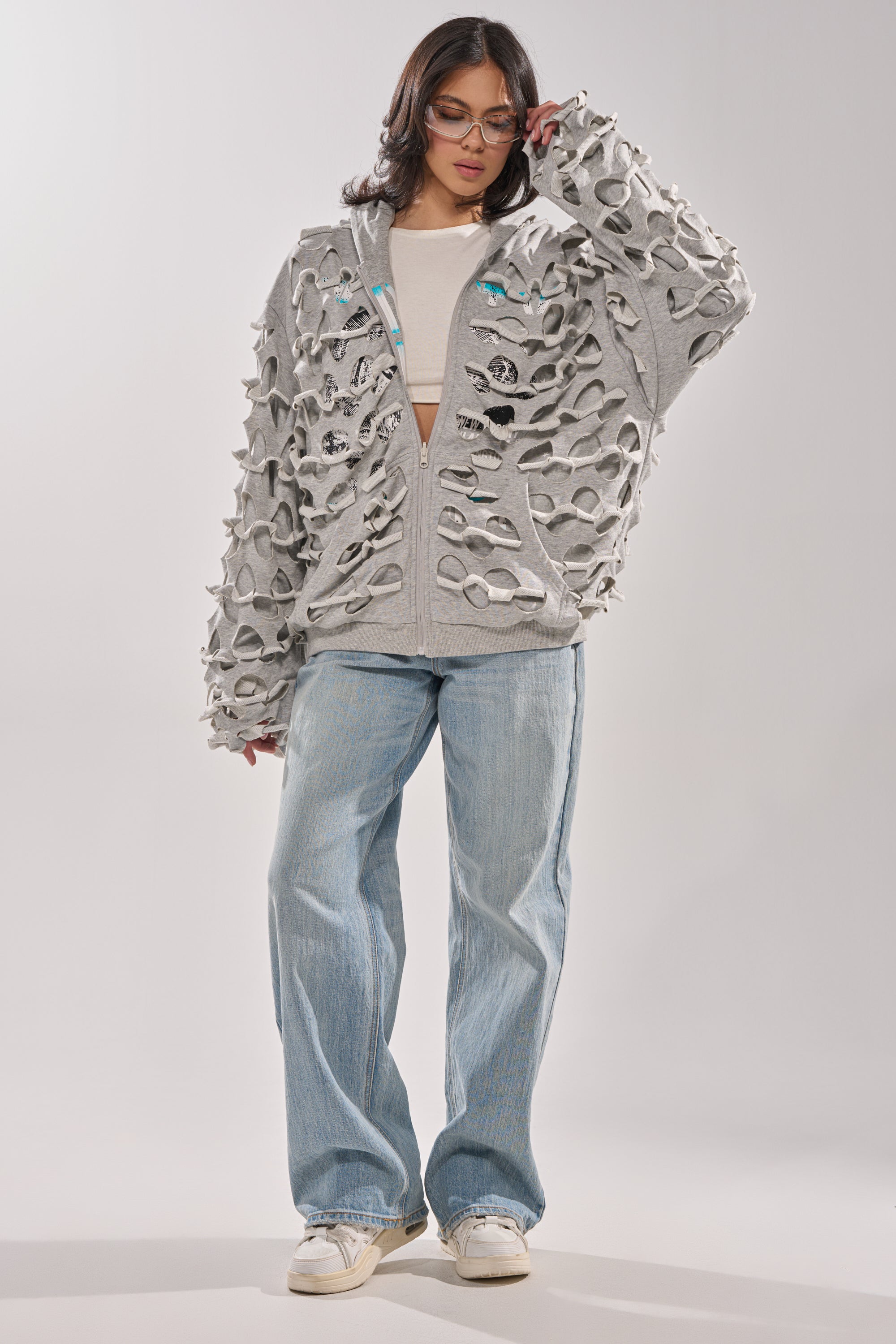 A person stands facing forward, wearing glasses, a beige crop top, the EPIC REVERSIBLE ZIP UP SWEATSHIRT with large cutouts, loose light blue jeans, and white sneakers against a plain light background. 14