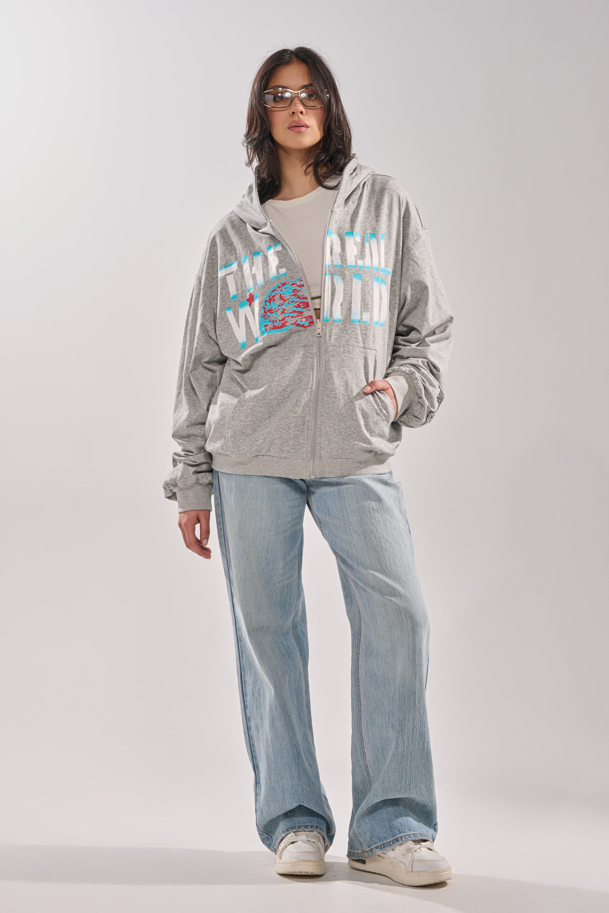 A woman wearing sunglasses, the EPIC REVERSIBLE ZIP UP SWEATSHIRT with colorful text, a white shirt, light blue wide-leg jeans, and white sneakers stands with one hand in her pocket against a plain background. 15