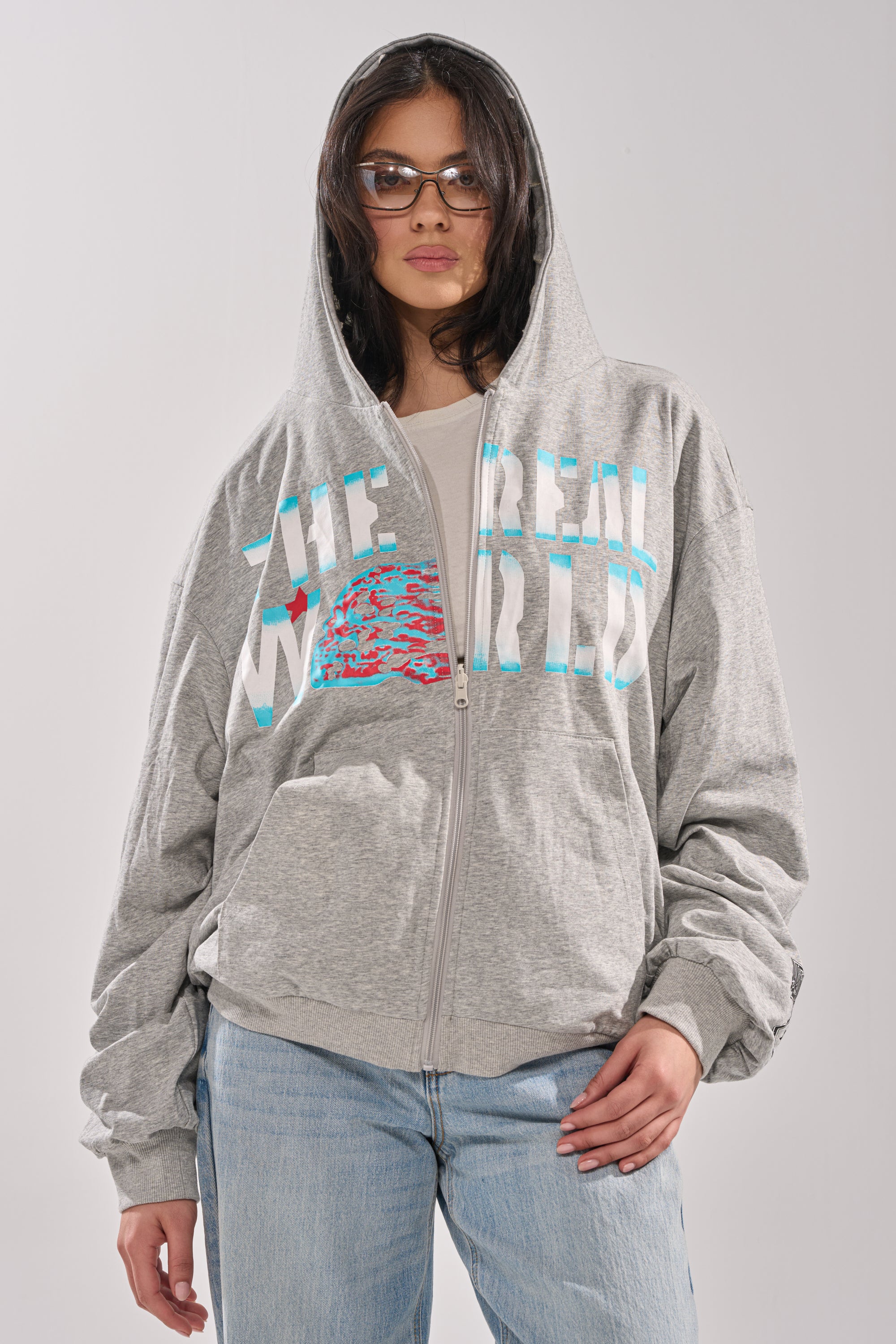 A person in glasses wears the EPIC REVERSIBLE ZIP UP SWEATSHIRT, light gray with "THE REAL WORLD" text and a colorful graphic, paired with light blue jeans and hood up, standing against a plain background. 1