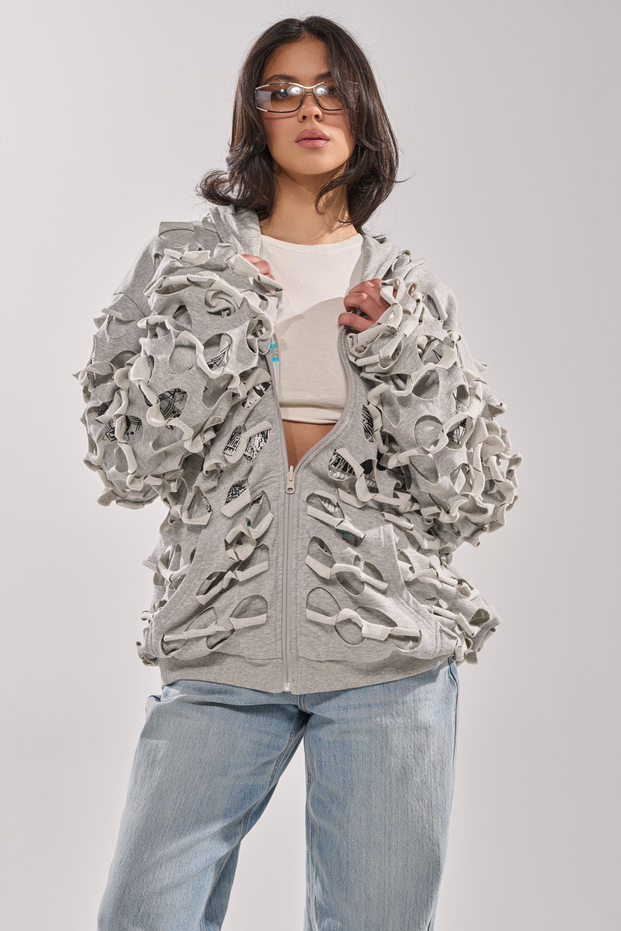 A woman in sunglasses, a white crop top, light blue jeans, and the EPIC REVERSIBLE ZIP UP SWEATSHIRT featuring unique textured cutout designs on the sleeves and front stands against a plain background. 2