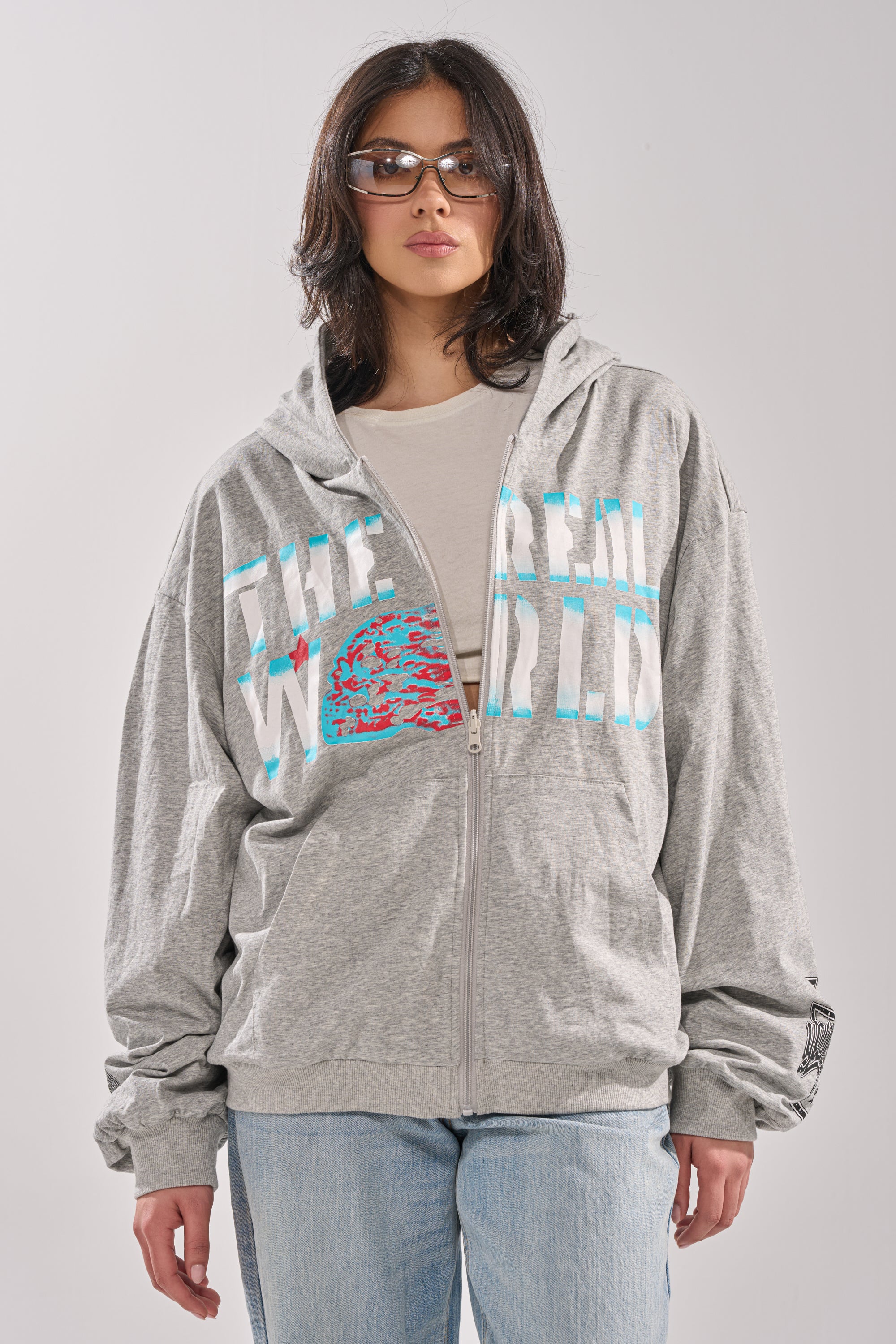 A person with shoulder-length dark hair wears sunglasses, the EPIC REVERSIBLE ZIP UP SWEATSHIRT in light gray with "THE REAL WORLD" text, a beige shirt underneath, and light blue jeans, standing against a plain light background. 7