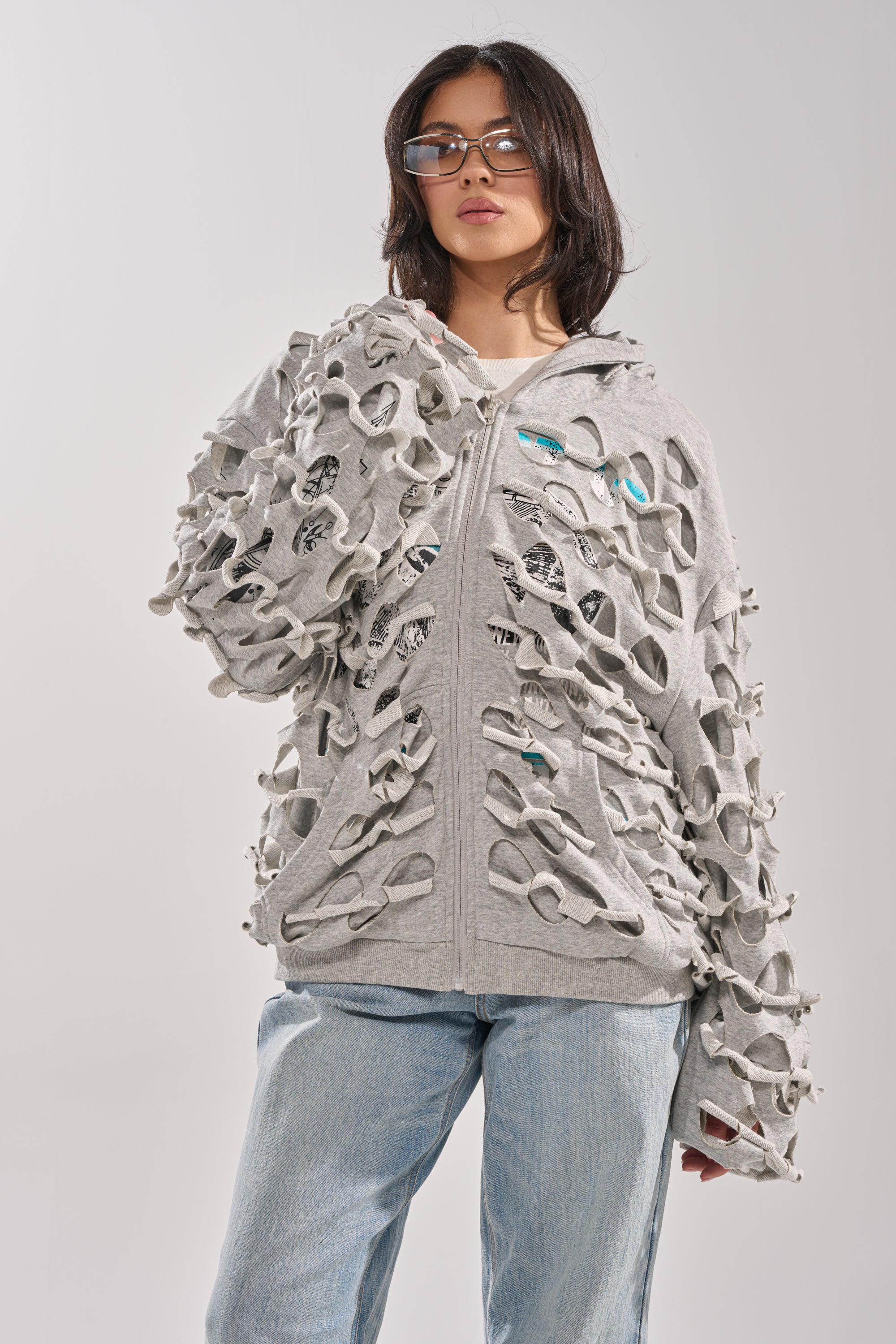 A woman with dark hair and glasses wears the EPIC REVERSIBLE ZIP UP SWEATSHIRT—grey, featuring large cut-out holes and an oversized hood—with light blue jeans, standing confidently against a plain, light background. 19
