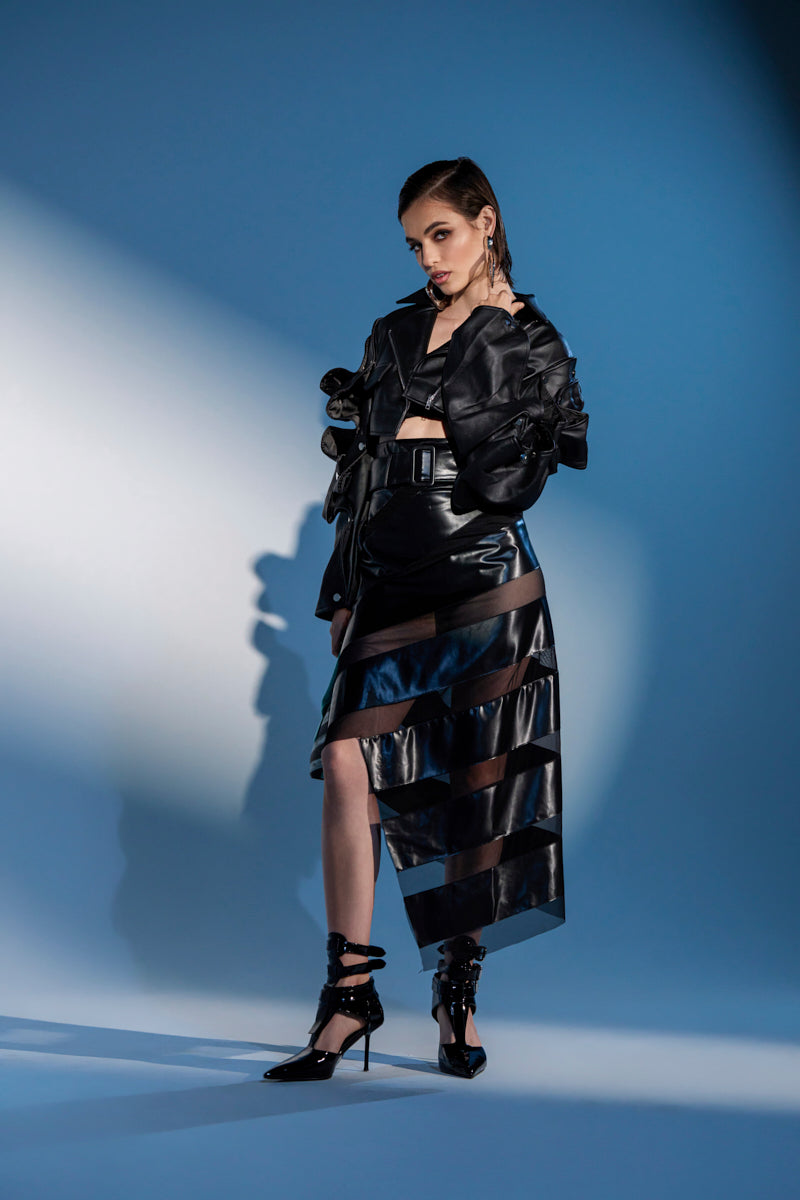 A woman poses confidently in a black leather jacket, the SOMETHING DIFFERENT MESH MIDI SKIRT WITH FAUX LEATHER featuring sheer horizontal panels, and black heels against a blue background with dramatic lighting.