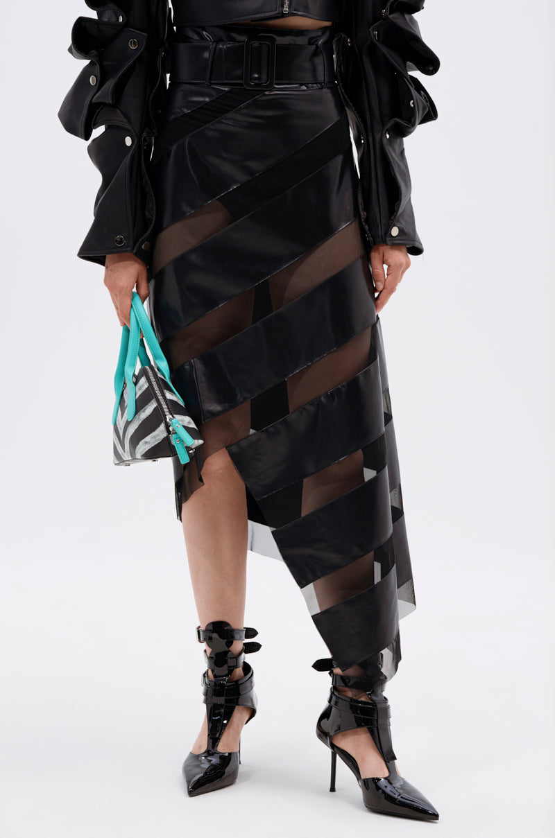 Wearing the SOMETHING DIFFERENT Mesh Midi Skirt with Faux Leather, a black jacket, black pointed heels with bows, and holding a small patterned clutch with turquoise handles.