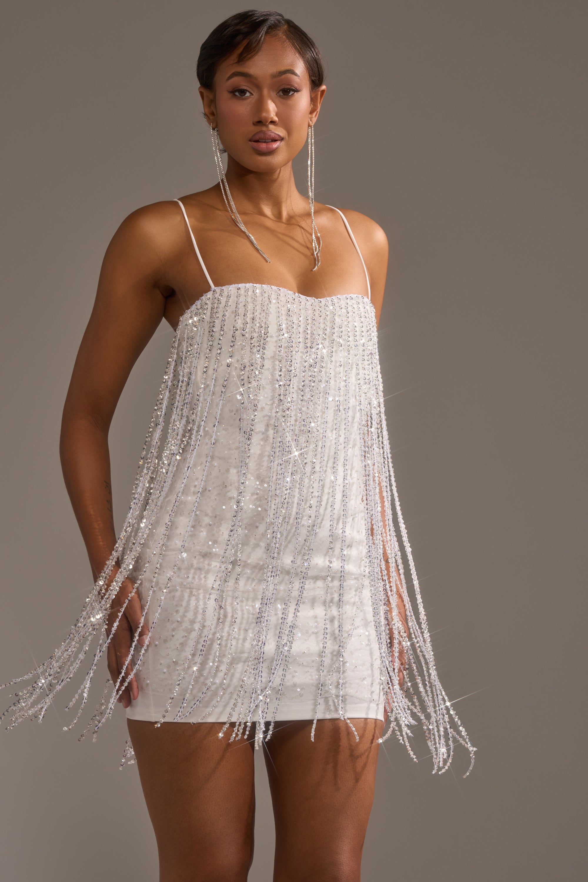 A woman stands confidently against a gray background wearing the CAME THRU DRIPPIN' FRINGE MINI DRESS, a white, beaded fringe mini dress with thin shoulder straps.