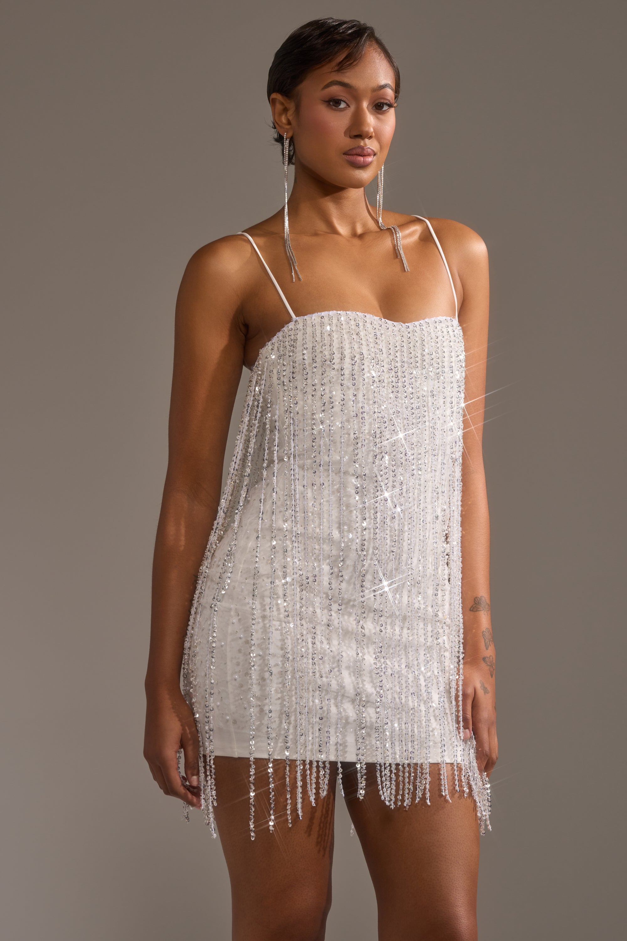 A woman models the CAME THRU DRIPPIN' FRINGE MINI DRESS, featuring sparkly silver beaded fringe and thin straps. She pairs it with long dangling earrings and poses against a plain gray background.