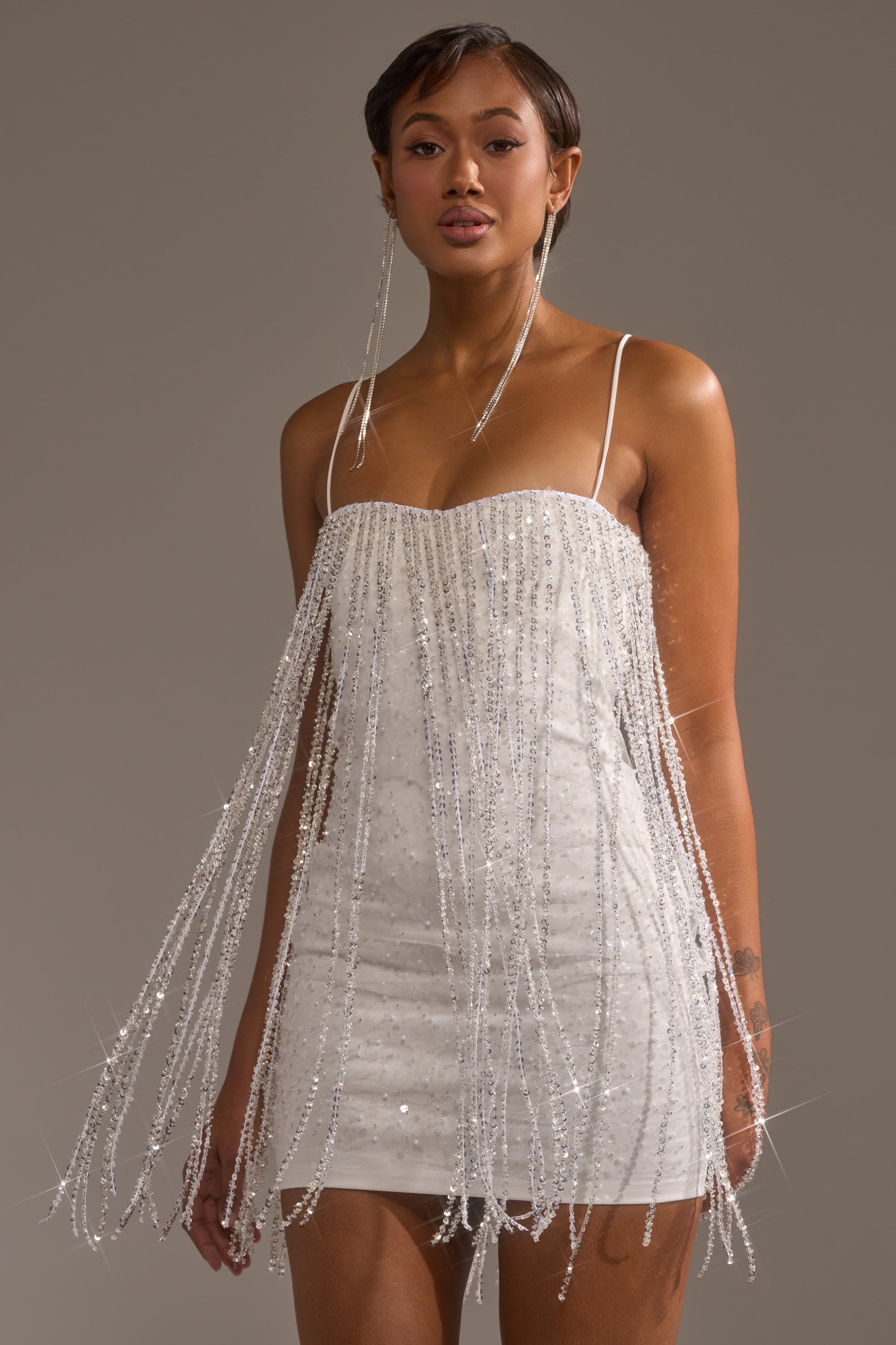 A woman poses elegantly against a plain gray background in the CAME THRU DRIPPIN' FRINGE MINI DRESS, featuring thin straps and long beaded fringe strands for a stunning, standout look.