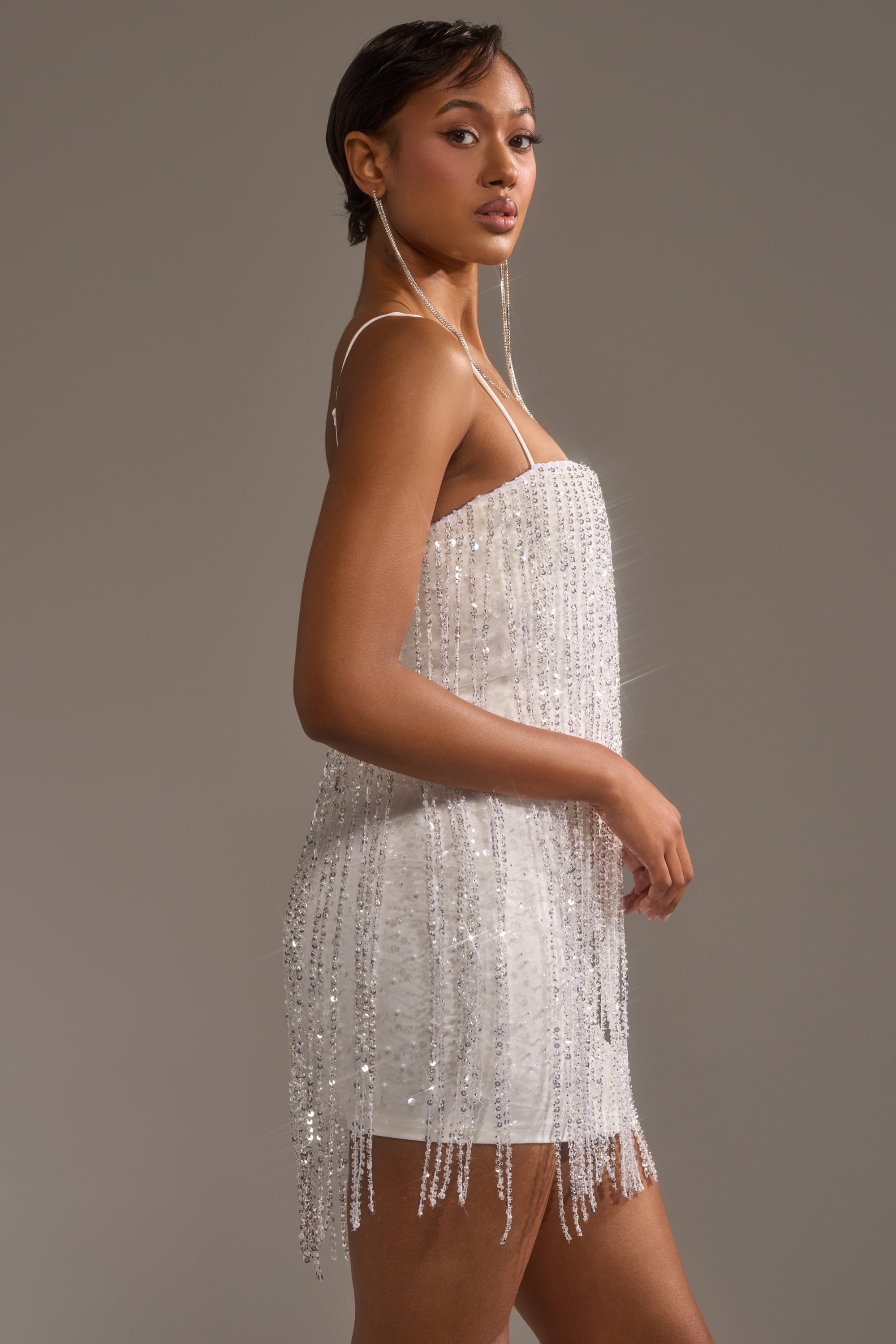 A woman poses confidently in the CAME THRU DRIPPIN' FRINGE MINI DRESS, a fitted, sleeveless beaded mini sparkling with fringe details, against a plain gray background with one hand by her side.