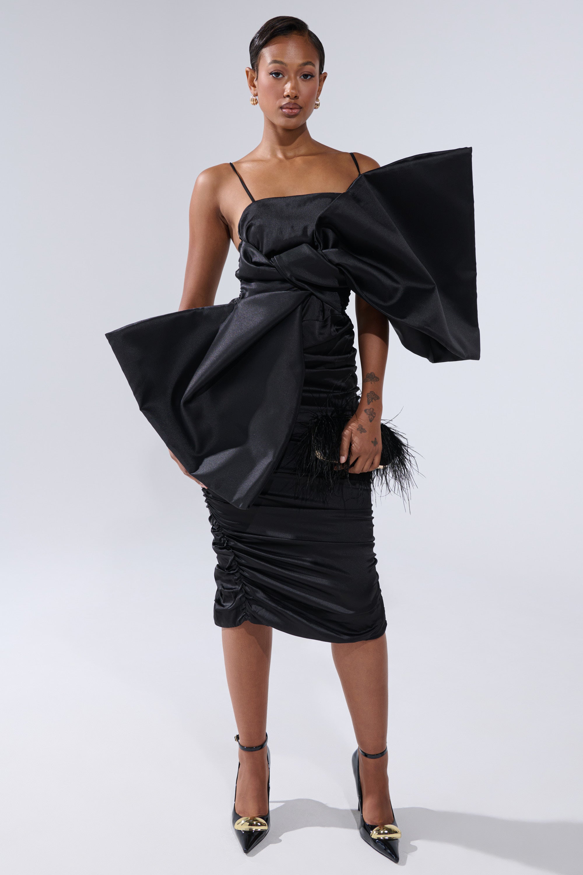 A woman stands confidently in the ONE OF ONE BOW MIDI DRESS IN BLACK, featuring an oversized bow on one shoulder. She completes the look with a black feather-accented clutch and black heels with gold tips against a plain light background.