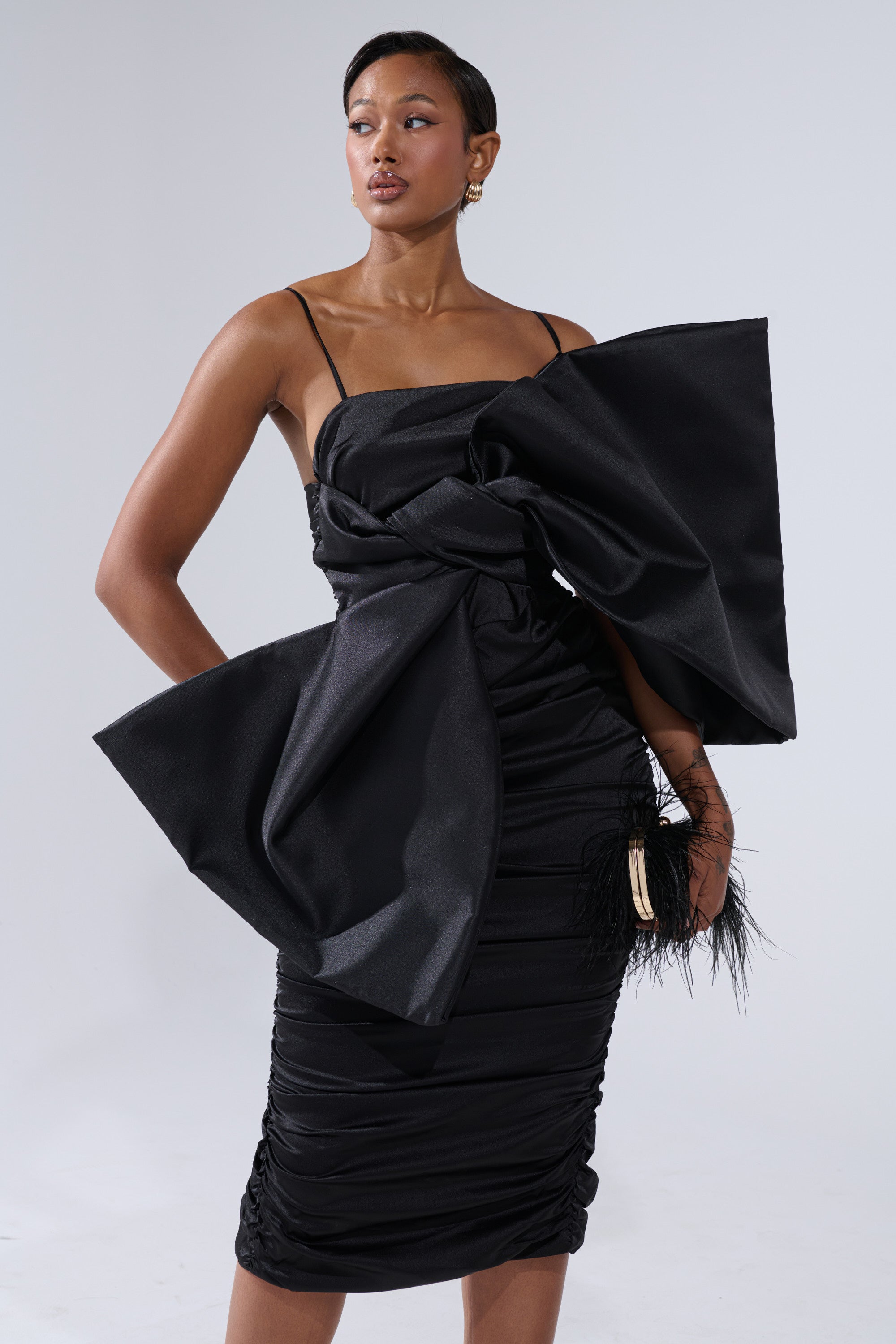 A woman poses in the ONE OF ONE BOW MIDI DRESS IN BLACK, a fitted satin dress with thin straps, dramatic front bow detail, ruched sides, and feather accents near the waist, set against a plain light gray background.