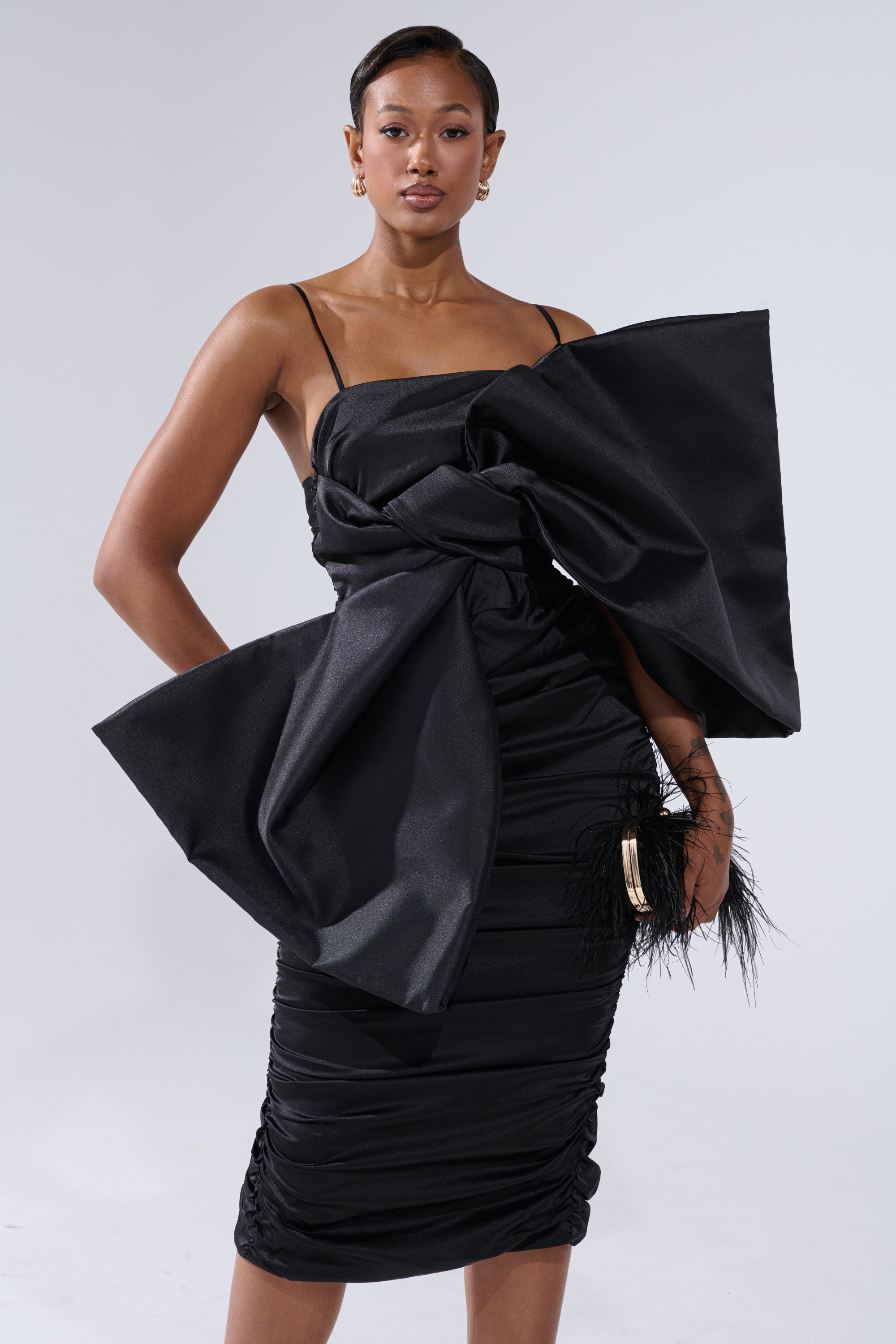 A woman stands confidently in the ONE OF ONE BOW MIDI DRESS IN BLACK, featuring a fitted silhouette and thin straps, as she holds a black clutch with feather accents against a plain light background.