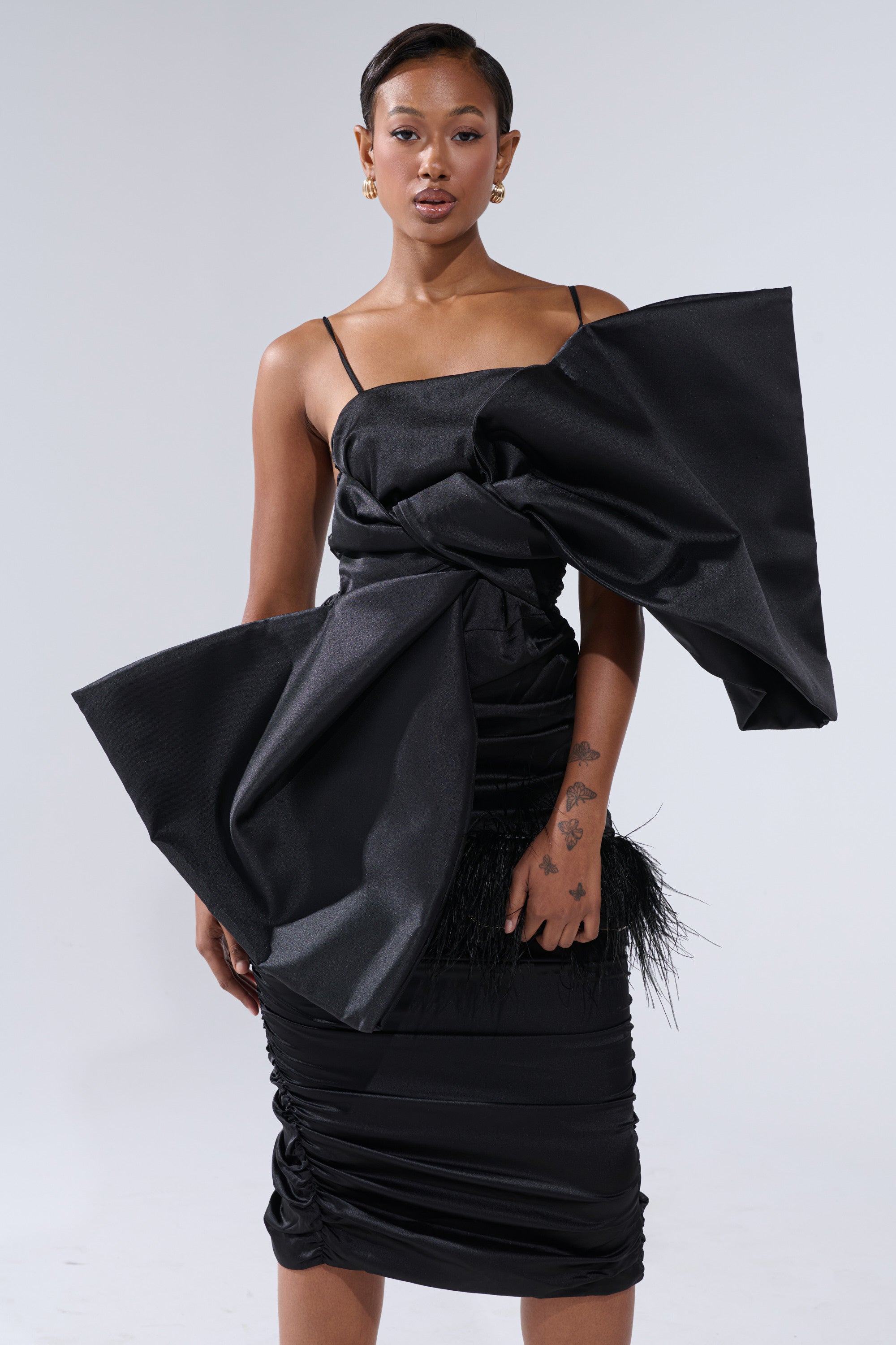 A woman with short hair, gold earrings, and tattoos on her left hand and arm poses against a plain white background in the ONE OF ONE BOW MIDI DRESS IN BLACK—a chic satin midi dress with thin straps and a dramatic oversized front bow.