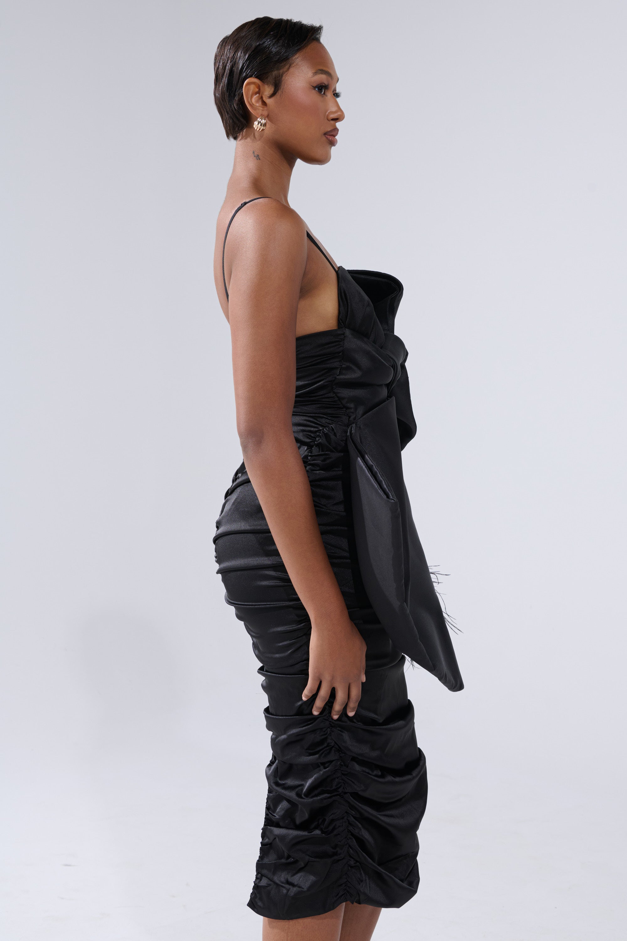 A woman with short hair stands in profile, showcasing the ONE OF ONE BOW MIDI DRESS IN BLACK—a fitted satin dress with thin straps and a large side bow—paired with elegant earrings against a plain light background.