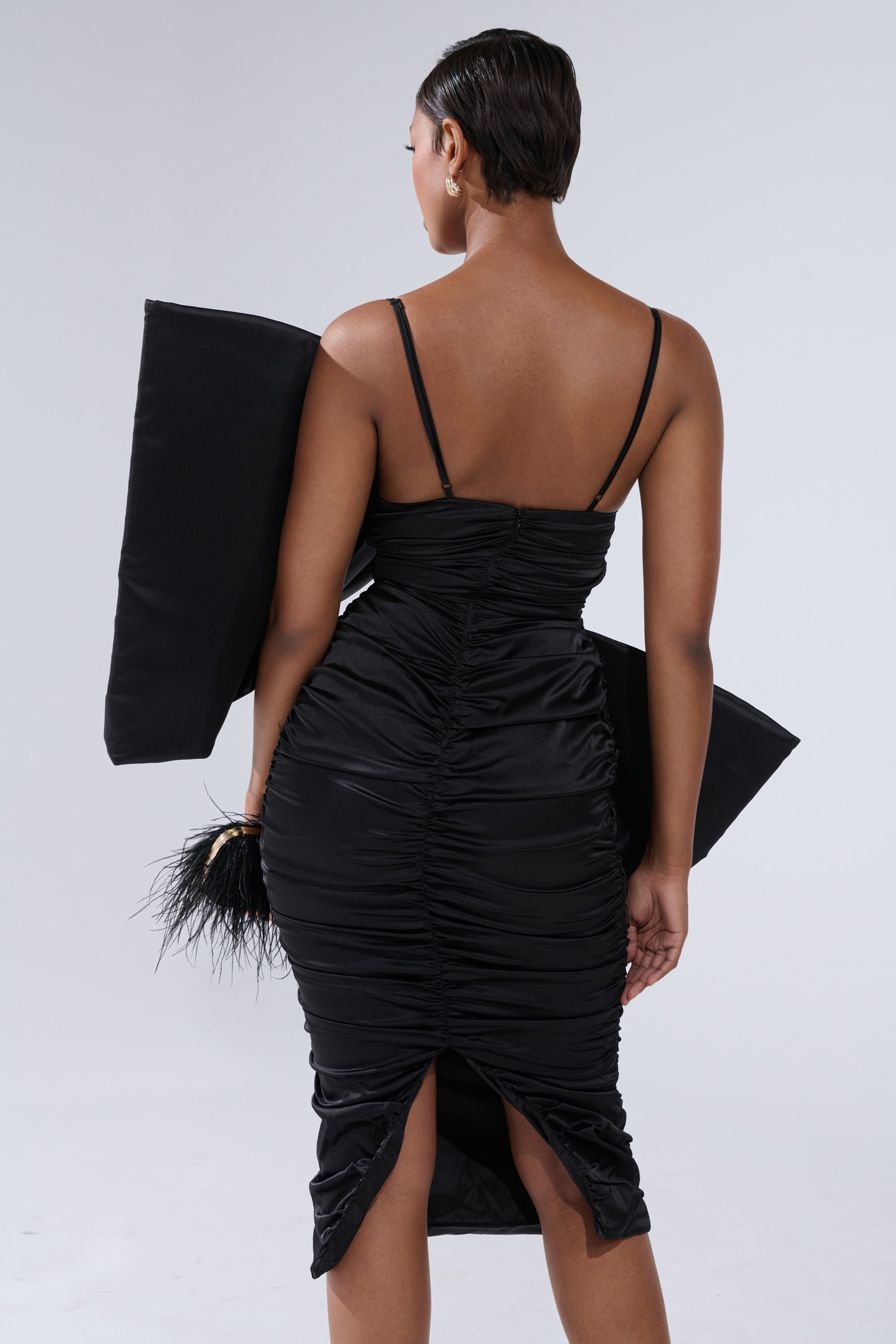 A woman wears the ONE OF ONE BOW MIDI DRESS IN BLACK, featuring spaghetti straps and a ruched bodycon fit, standing with her back to the camera while holding a large black rectangular item and a feathered black accessory.