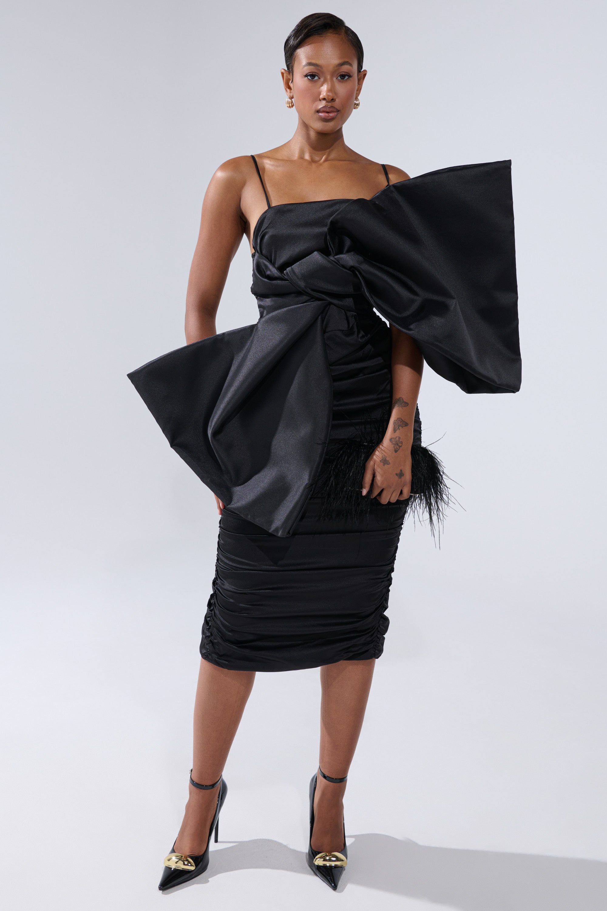 A woman exudes confidence in the ONE OF ONE BOW MIDI DRESS IN BLACK, featuring thin straps and an oversized bow detail, styled with black heels and a sleek clutch.