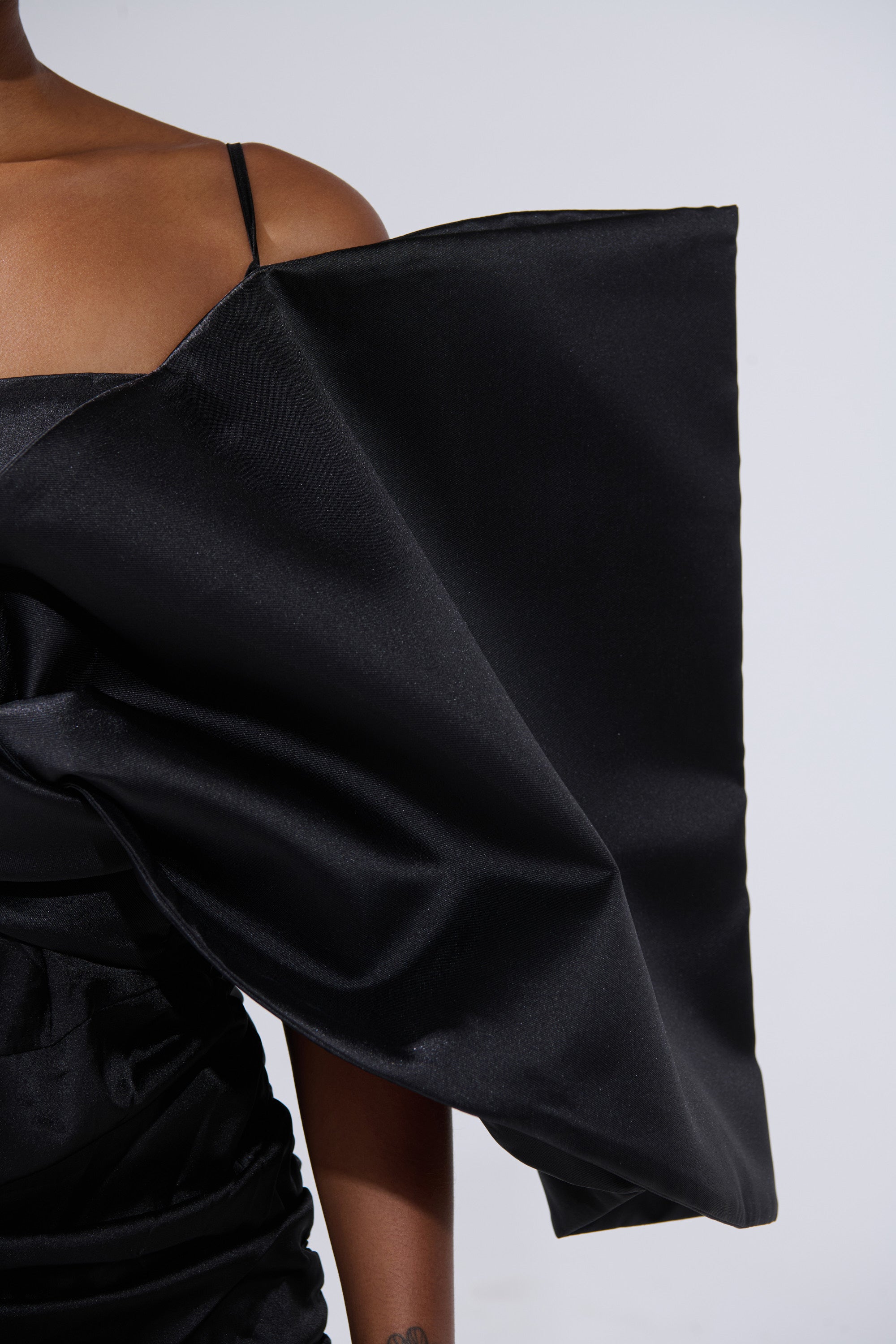 A person wears the ONE OF ONE BOW MIDI DRESS IN BLACK, featuring a large, structured angular bow detail on one shoulder. The plain, light background highlights the striking upper half of this bodycon LBD.