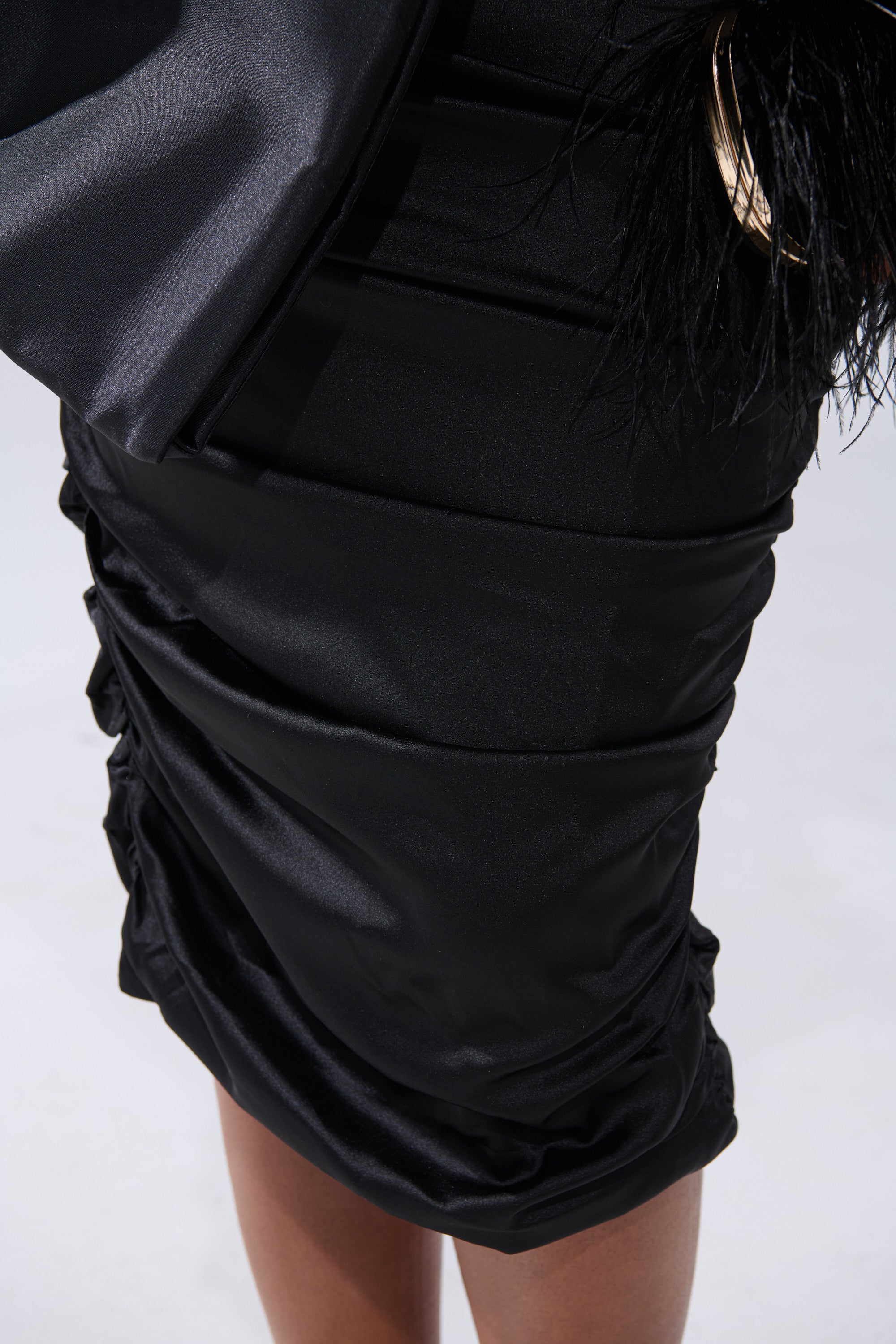 A close-up of a person wearing the ONE OF ONE BOW MIDI DRESS IN BLACK, featuring black satin ruching, a hint of a black feathered accessory, and their dark-skinned legs against a light background.