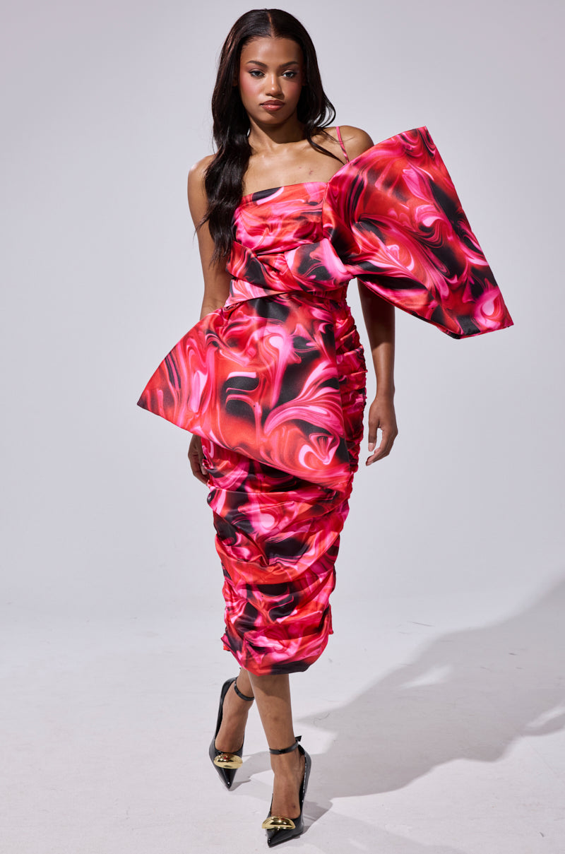 A woman models the ONE OF ONE BOW MIDI DRESS, featuring a ruched pink and black design with an asymmetrical silhouette, oversized bow detail, spaghetti straps, and paired with black heels with gold embellishments.