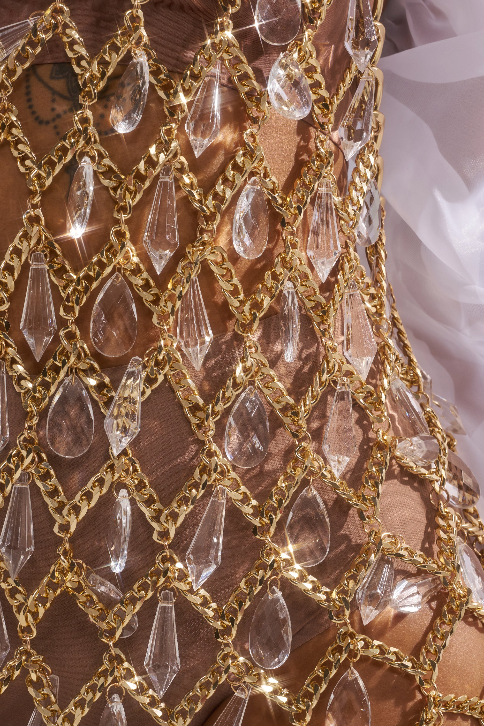 Close-up of the OH HOT DAMN RHINESTONE MINI DRESS featuring a gold chain mesh halter neckline embellished with clear crystal teardrop and prism pendants, draped over sheer nude fabric revealing a partial tattoo underneath.
